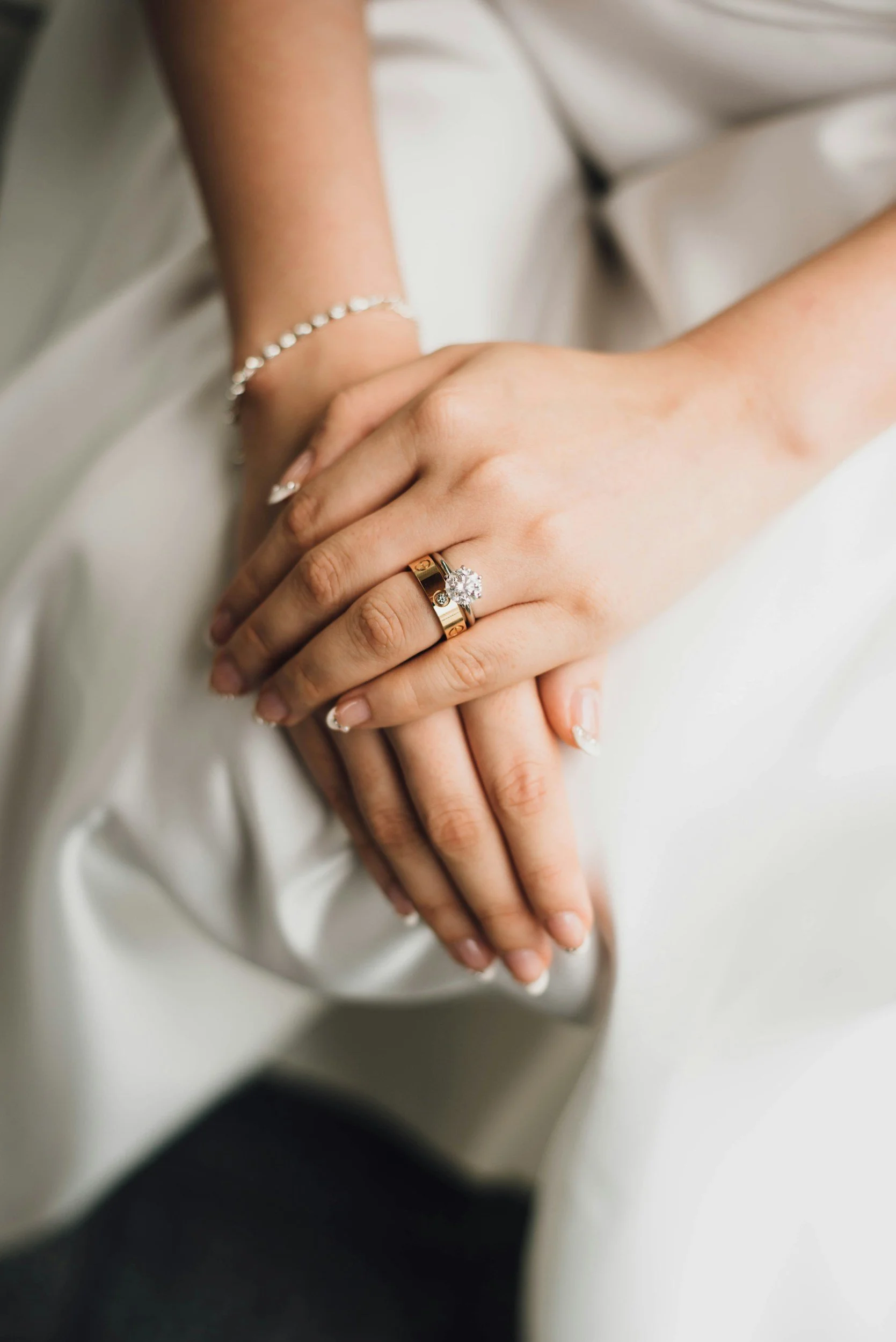 Close-up of a woman's hand wearing a wedding ring with a large diamond, resting on another person's hand with a bracelet and wedding band, against a white satin fabric background.