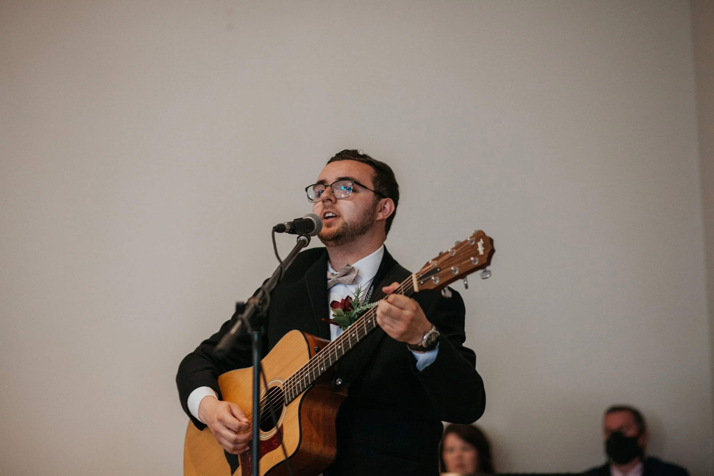 Man in a black suit and glasses playing an acoustic guitar and singing into a microphone at an event.