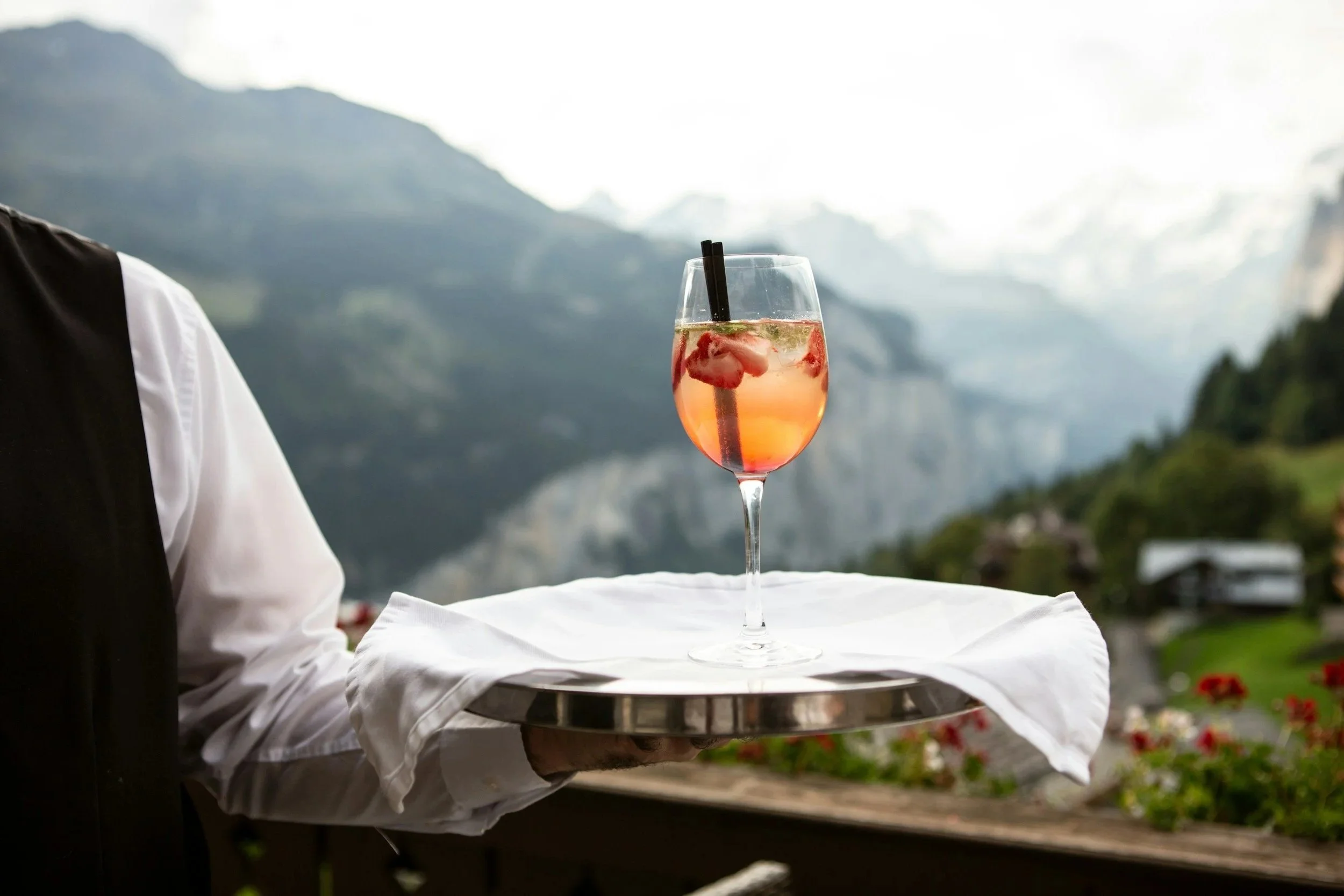 A server holds a tray with a glass of pink cocktail garnished with fruit, outdoors with a mountain landscape in the background.