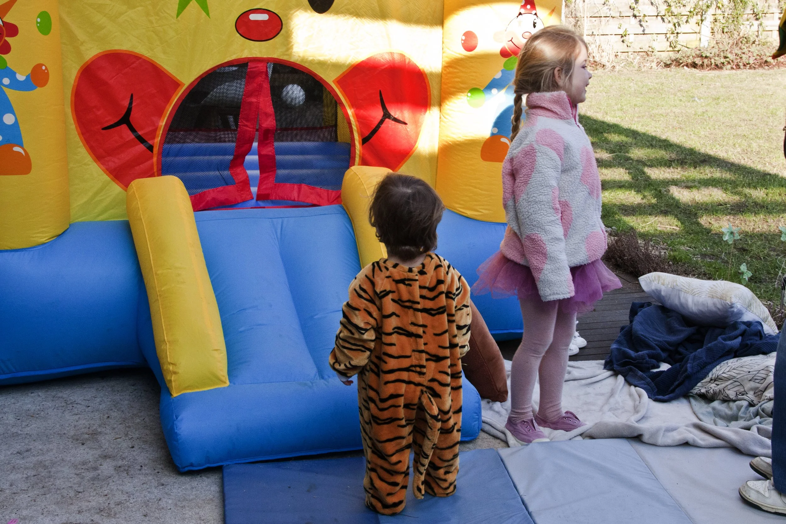 Twee kinderen staan voor een kleurrijk springkussen buiten, met een dekbed en kussens op de grond.
