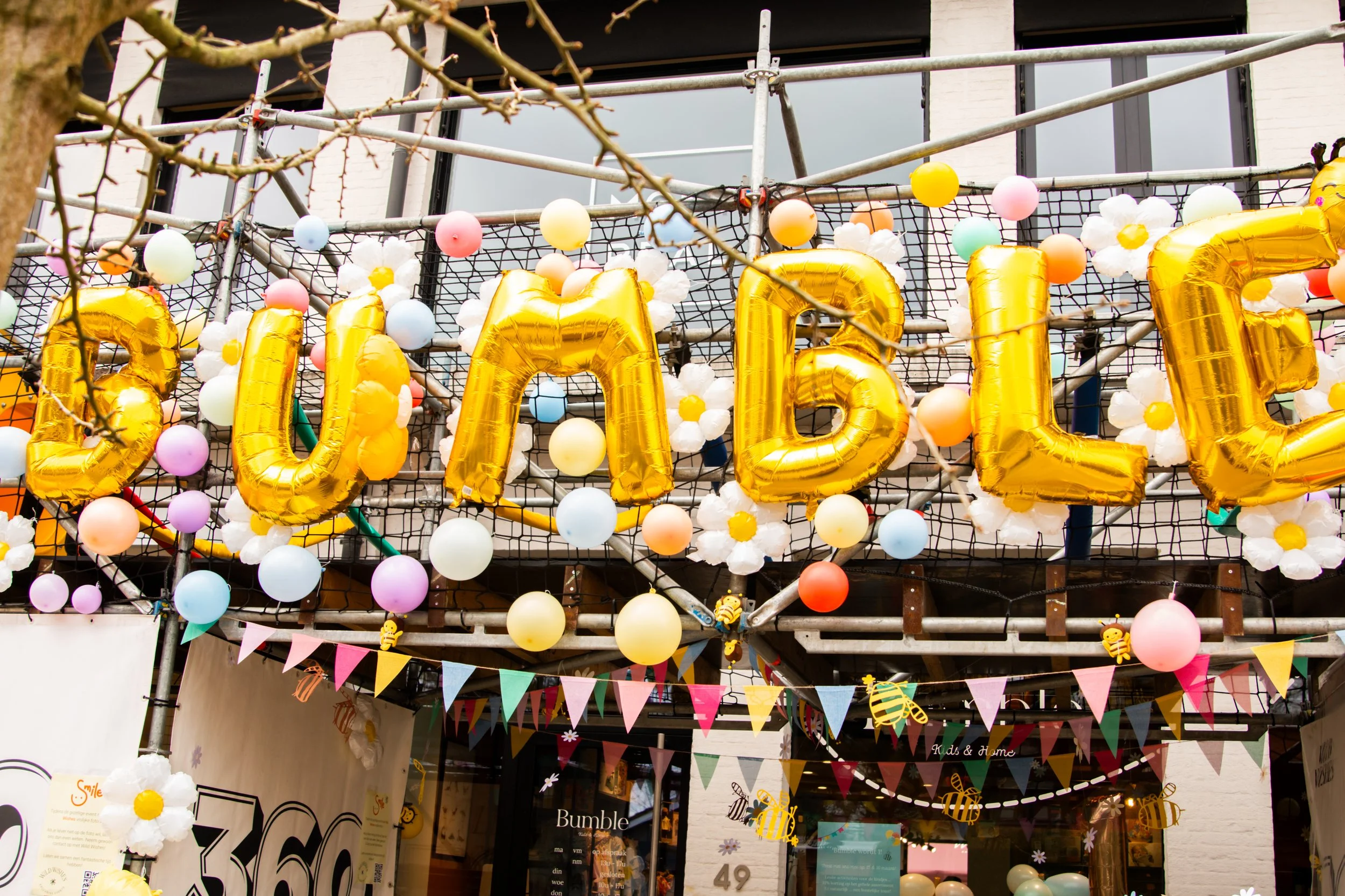Gouden ballonnen vormen het woord 'Bumble', omringd door kleurrijke ballonnen en slingers tegen een winkelgevel. Er zijn bloemen- en bijenversieringen aanwezig.