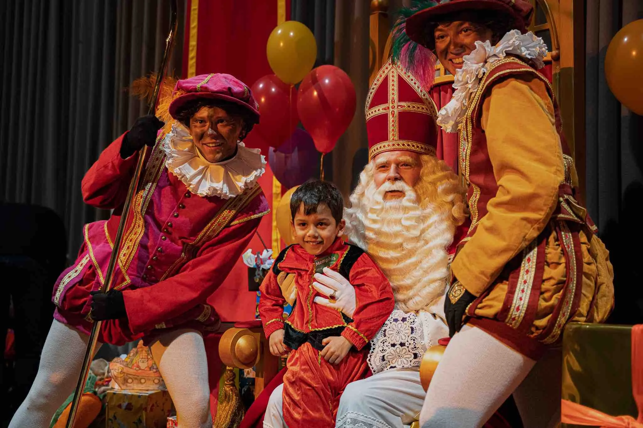 Een jongen zit op schoot bij Sinterklaas, omringd door drie verklede mensen in kleurrijke kostuums met zwarte schmink op hun gezichten, in een feestelijke setting met ballonnen op de achtergrond.