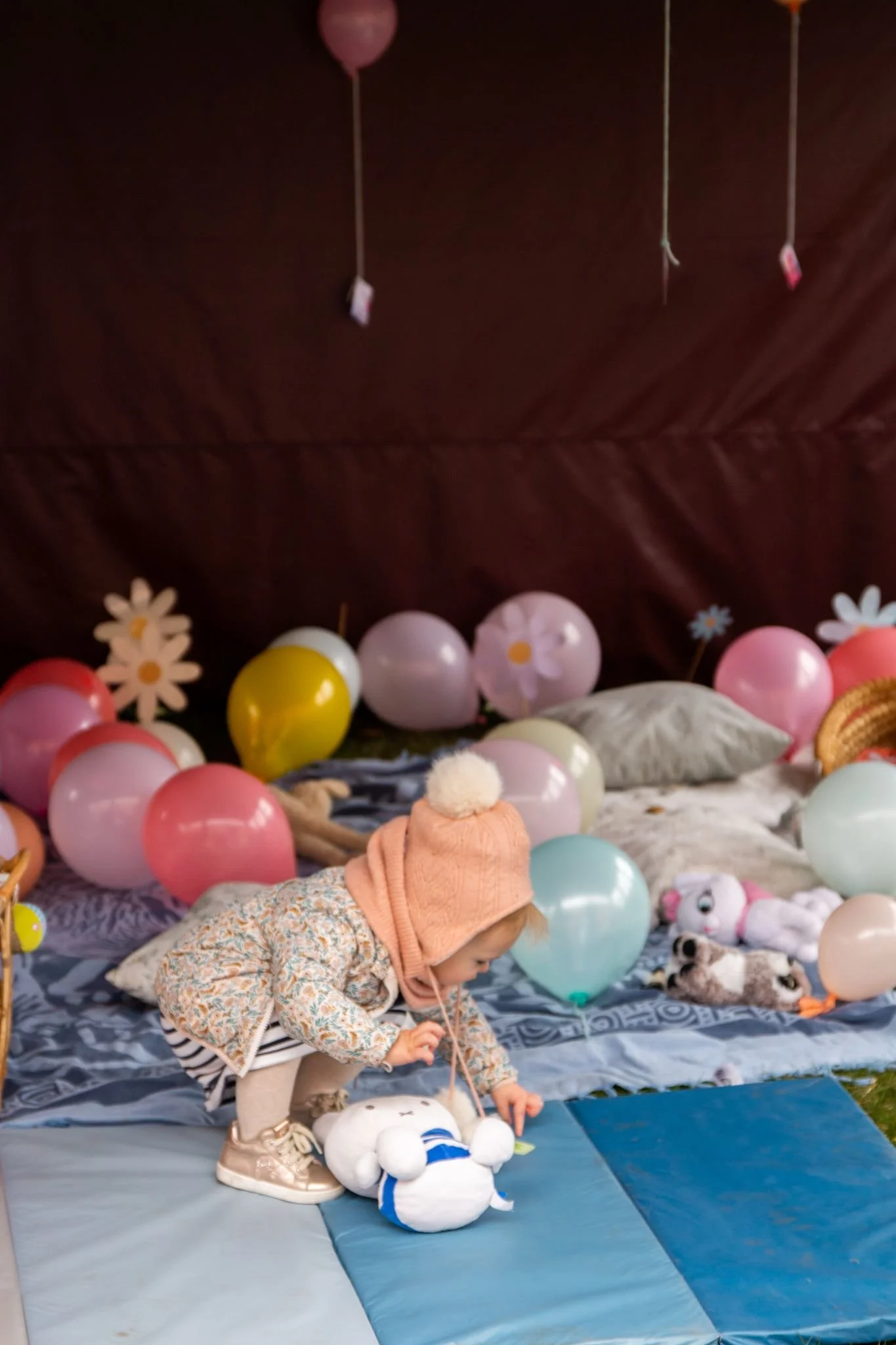 Een jong kind speelt met een knuffel op een blauwe mat, omringd door kleurrijke ballonnen in een feestelijke setting. Er zijn ook andere knuffels en kussens op de achtergrond.
