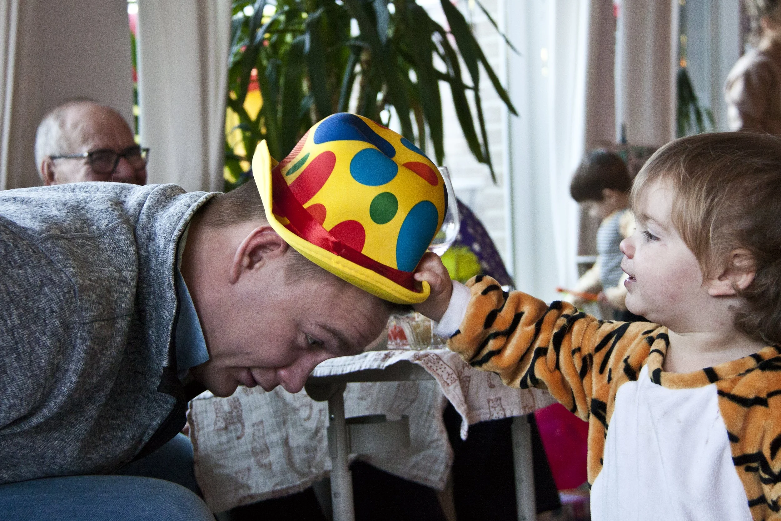 Een kind in een tijgerkostuum trekt aan de hoed van een man met een bontgekleurde hoed. Op de achtergrond zit een oudere man met een bril.