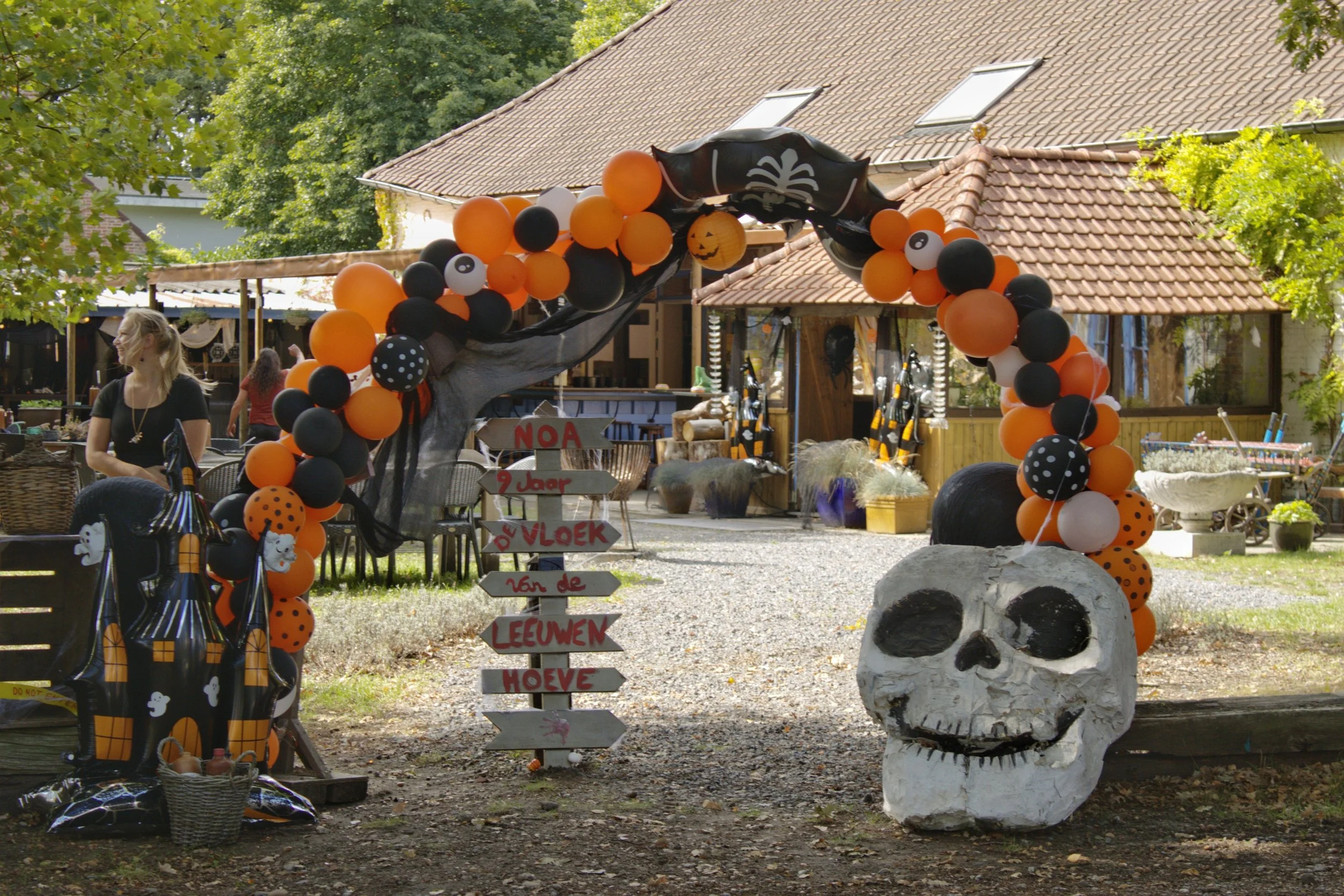 Halloween decoratie met een arendvormige ballonboog met zwart, oranje, witte en met stippen balonnen, een groot nepskelet, een groot zwarte pompoen, en een houten bord met Nederlandse tekst voor een feestje.