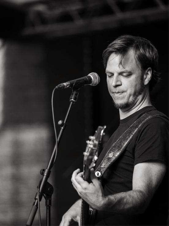Zwart-wit foto van een man die basgitaar speelt op een podium met een microfoon dichtbij.