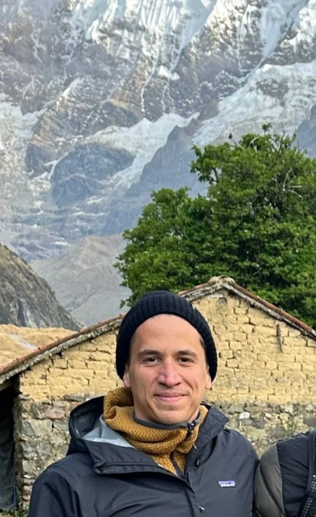 Person in winter clothing standing outdoors with a mountainous snowy background and stone building.