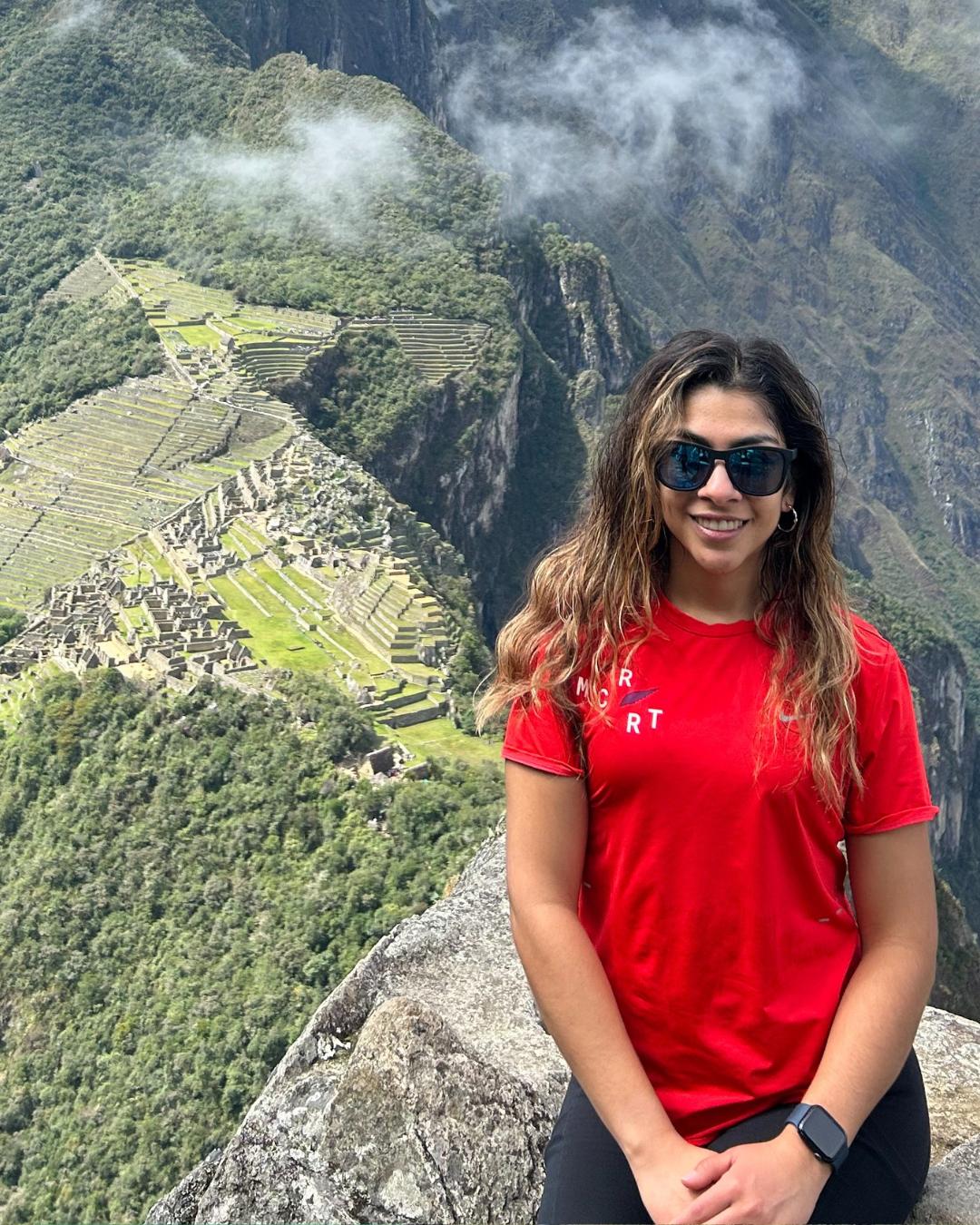 Person in winter clothing standing outdoors with a mountainous snowy background and stone building.