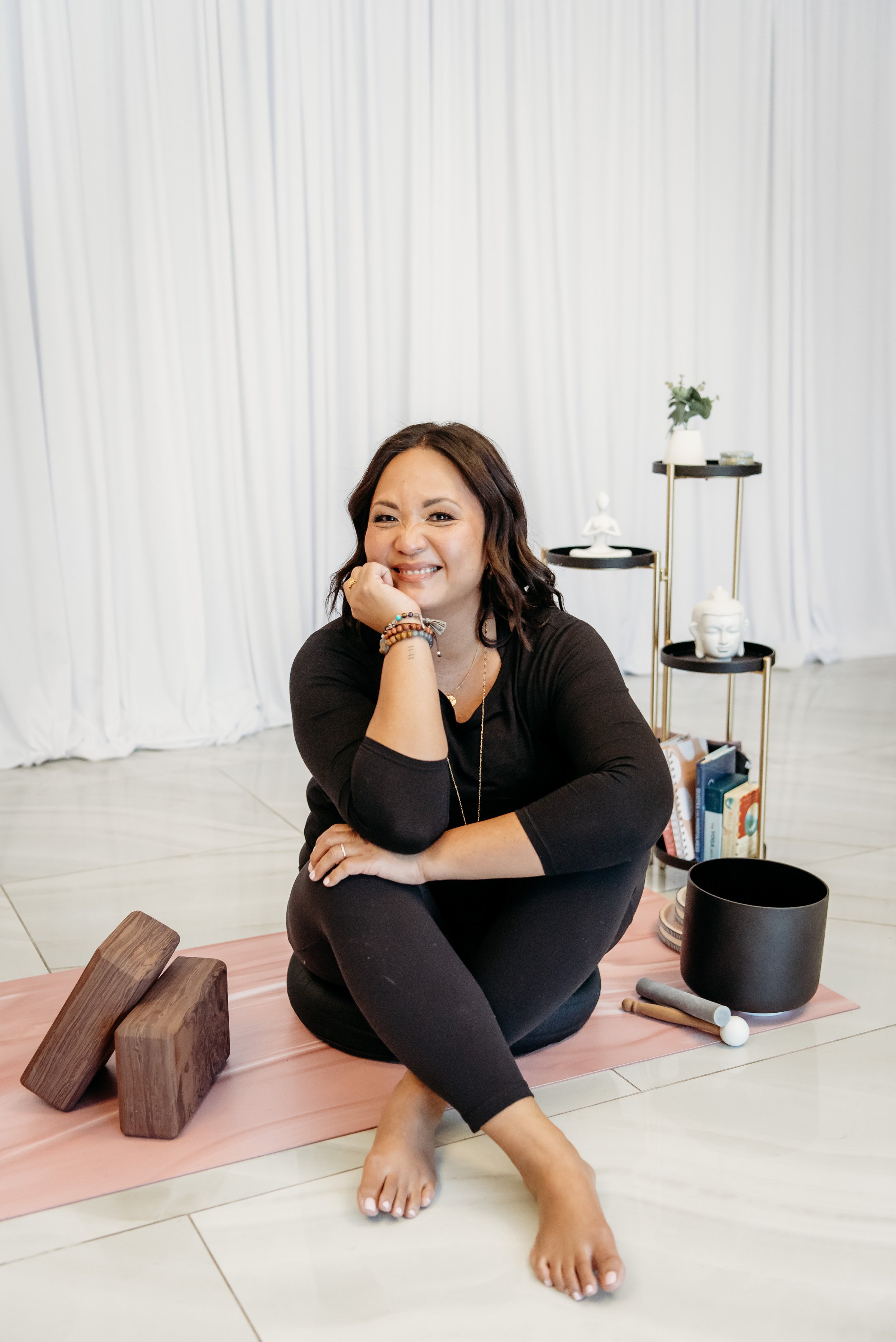 JoAnn Drasdis practicing yoga sitting in a wide-legged stretch on a mat with elbows resting on two blocks, smiling, with a decorative wall pattern in the background.