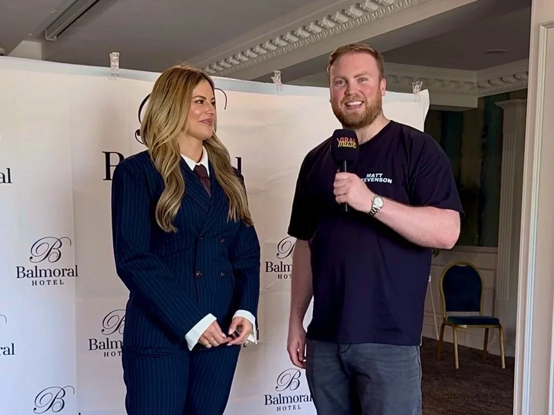 A man with a beard holding a microphone labeled 'VIBR Music' stands next to a woman in a blue pinstripe suit at a promotional event at the Balmoral Hotel. The woman has long blonde hair and is smiling, while the man is smiling and looking at the camera.
