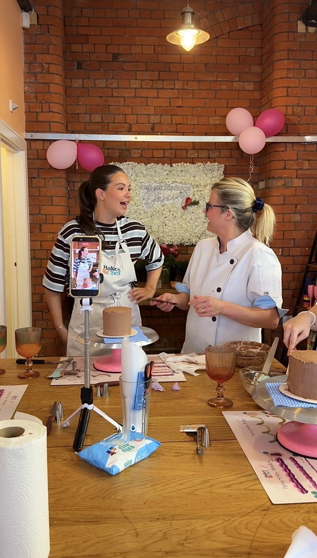 Two women, one young and one older, are having a conversation in a kitchen-style room with red brick walls. The young woman wears a striped shirt and apron, and the older woman wears a chef's coat. On the table are various baking tools, a chocolate cake, and ingredients. Pink and light pink balloons are decorated on the wall behind them, and a flower display is also in the background.