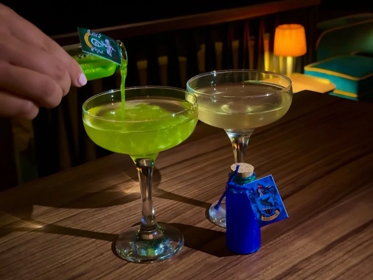 A person pouring green liquid into a cocktail glass, with a second cocktail glass and a small blue bottle with a cork and a label on a wooden table, dimly lit with a lamp and furniture in the background.