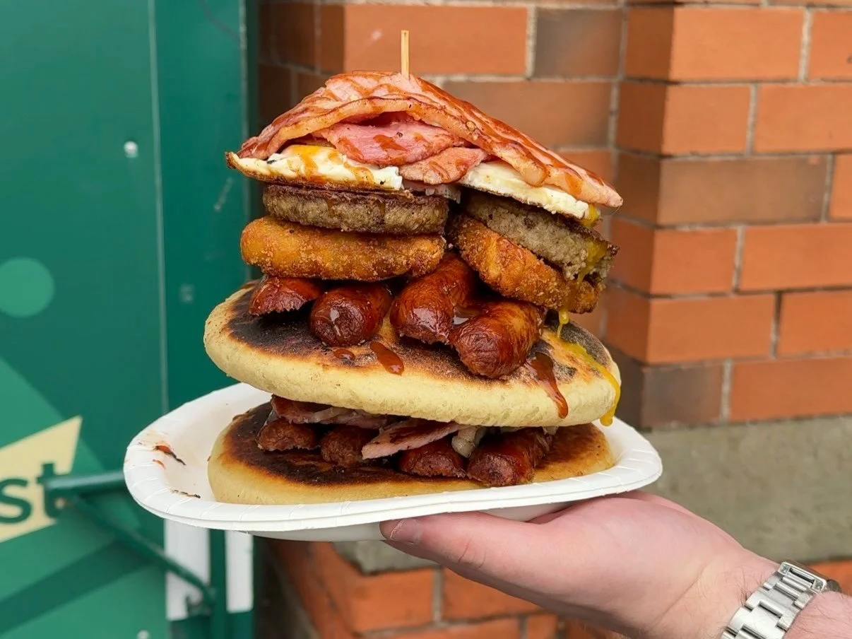 A hand holding a tray with an oversized burger layered with various fried foods, meats, and bread, against a brick wall background.