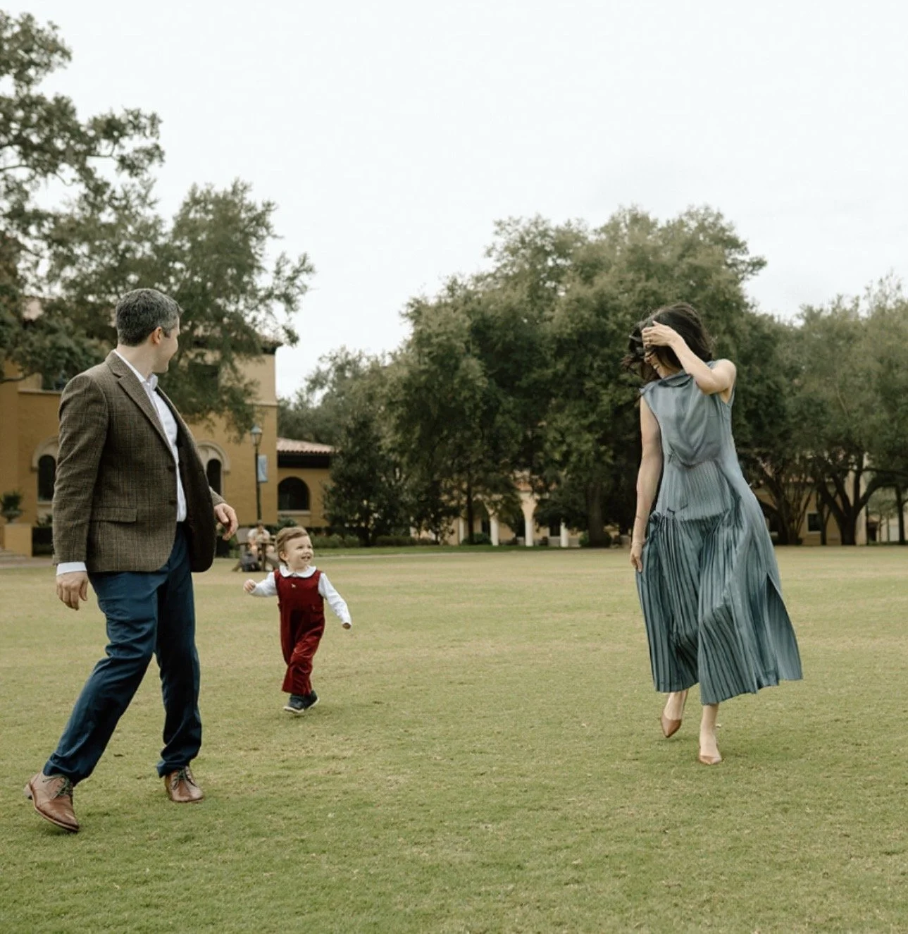 A family of three enjoying a day outdoors in a park. A man and a woman are running with a young boy between them, all smiling and looking happy. The woman is wearing a flowing dress, and the man is dressed in a blazer and pants. The park has green grass and trees, with a building in the background.