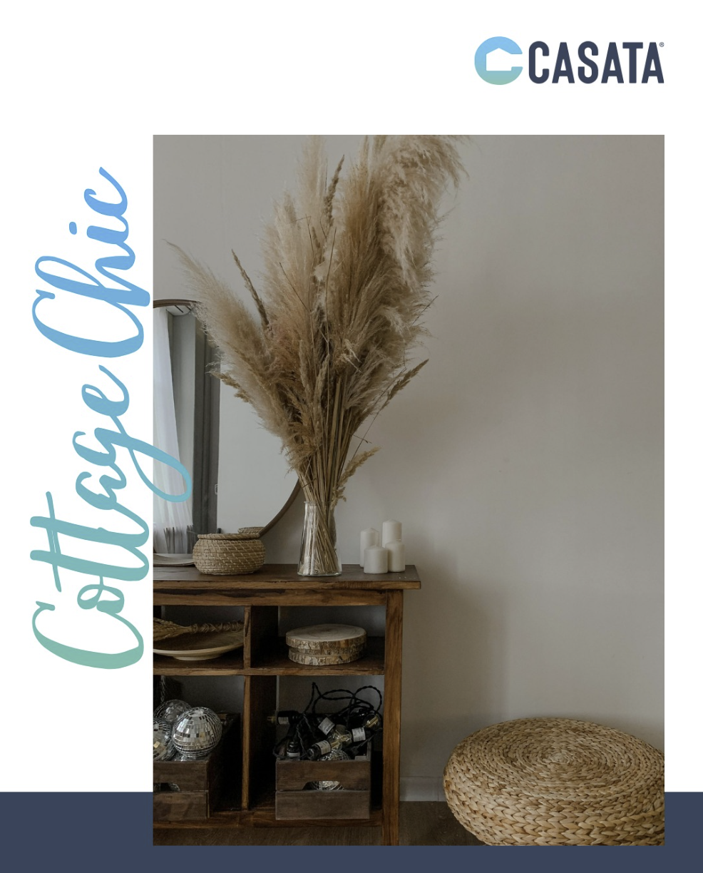 Casata instagram post A wooden console table with a mirror, decorative baskets, white candles, and a large vase of pampas grass against a plain wall, with a woven round pouf nearby.