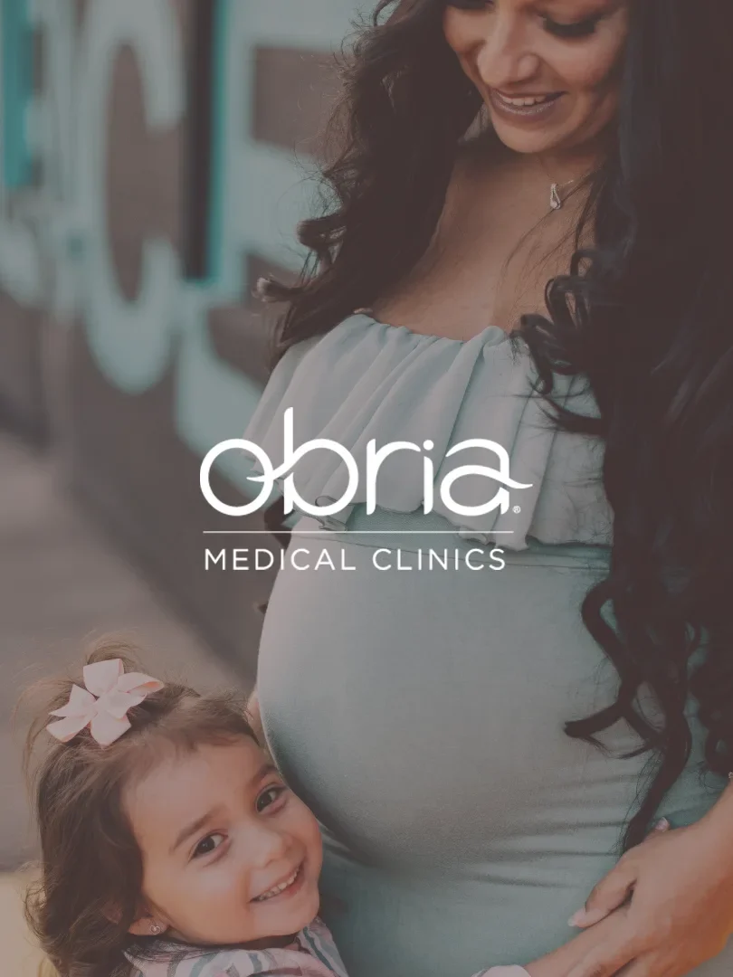 A pregnant woman with long dark curly hair smiling and standing outdoors, with a young girl with light brown hair and a pink bow smiling and hugging her. There is a logo overlay for "Obria Medical Clinics"