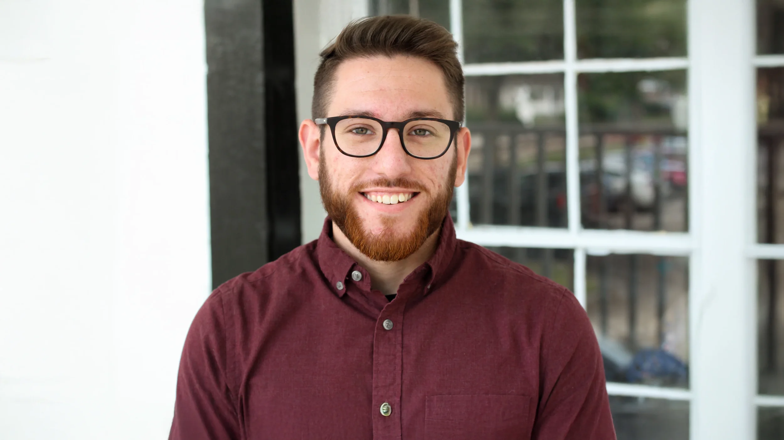 A young man with glasses, dark brown hair, and a beard smiling, wearing a maroon button-up shirt, standing outdoors near a building with large glass windows.