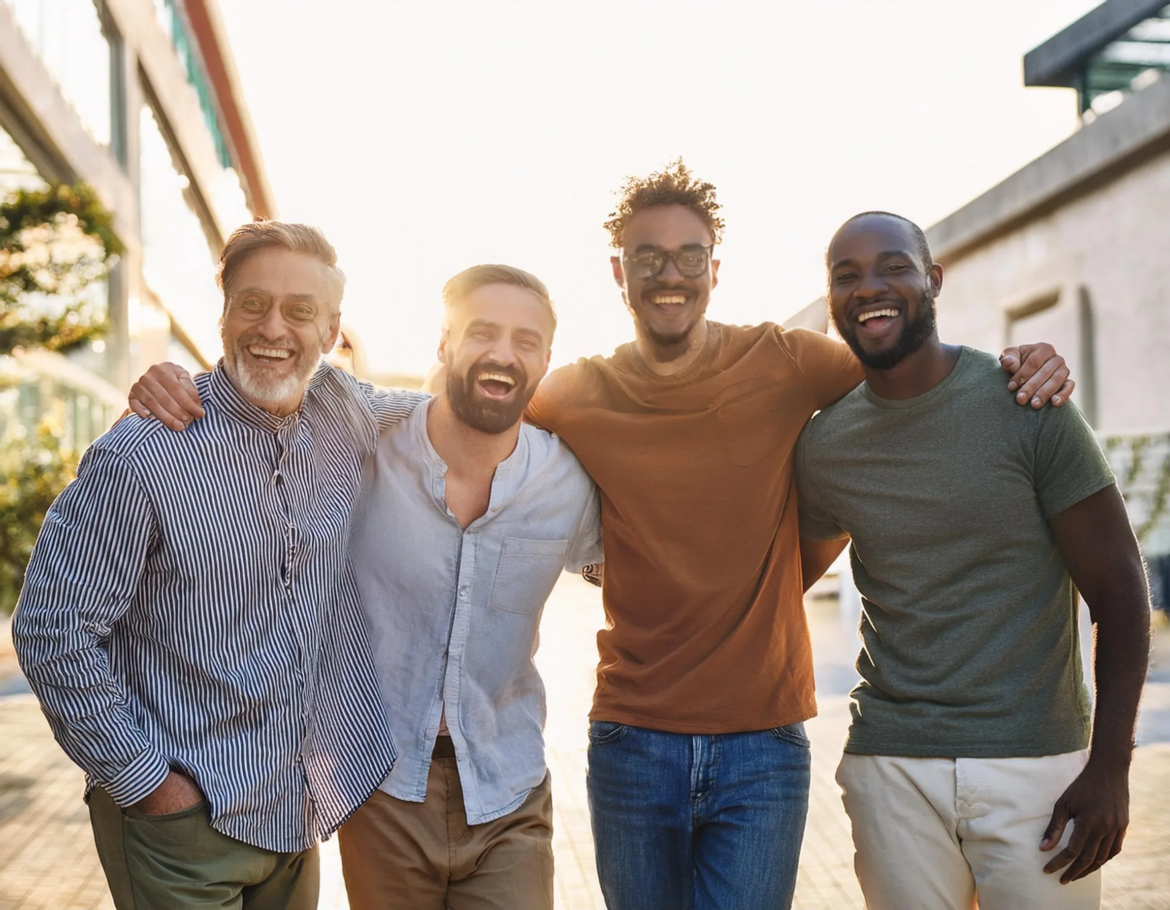 Four men are standing outdoors, smiling, with their arms around each other, enjoying a sunny day.