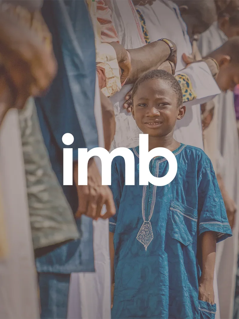A young boy in traditional African clothing standing among adults in colorful outfits, participating in an outdoor event or gathering. There is a logo overlay for "International Mission Board" with "imb" in lowercase letters.