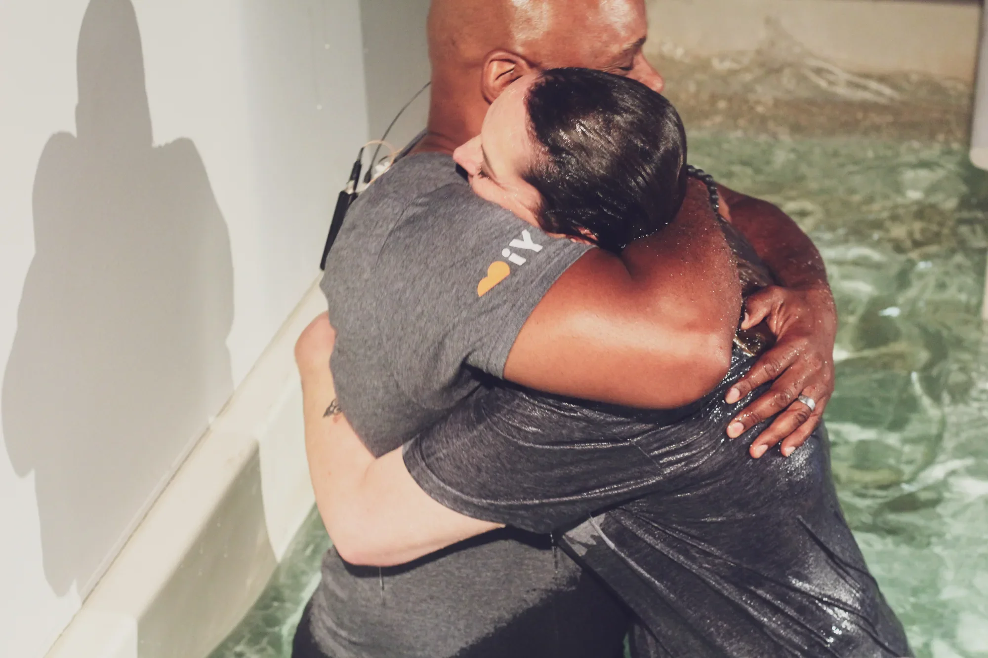 Two people are hugging in a baptism pool, water dripping from their bodies. One person's face is visible with eyes closed, and the other person's face is hidden. They are embracing each other tightly.