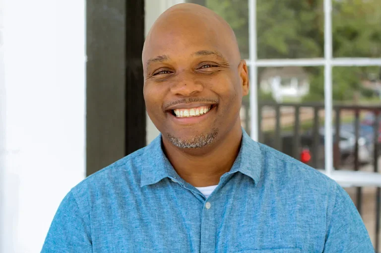 Smiling man in a blue shirt standing outdoors near a window.
