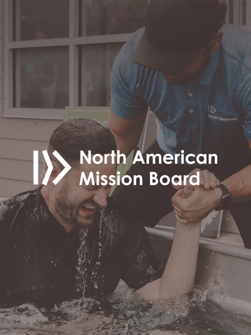 A man in water being baptized by another man outdoors, with a building and window in the background. There is a logo overlay for "North American Mission Board".