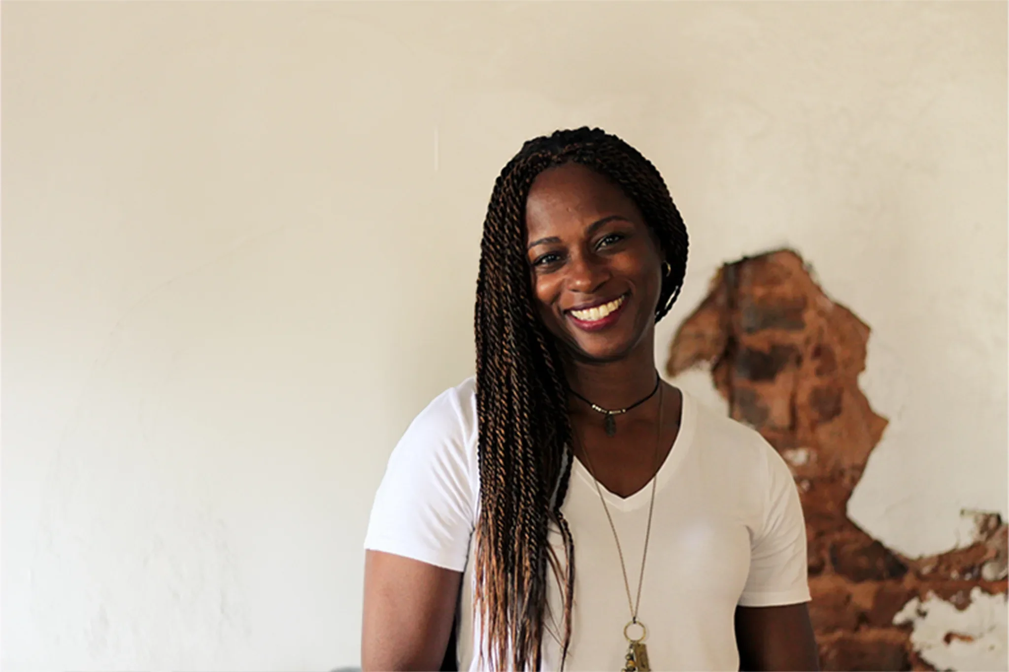 A smiling woman with long braided hair wearing a white T-shirt, necklace, and choker, standing indoors with a decorative wall piece in the background.