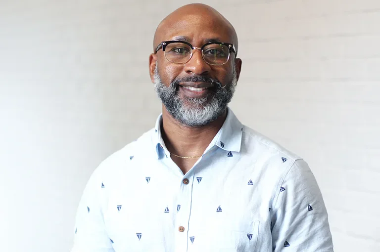 Portrait of a smiling Black man with glasses, a gray beard, and a shaved head, wearing a white button-up shirt with small sailboat patterns, standing against a plain white wall background.