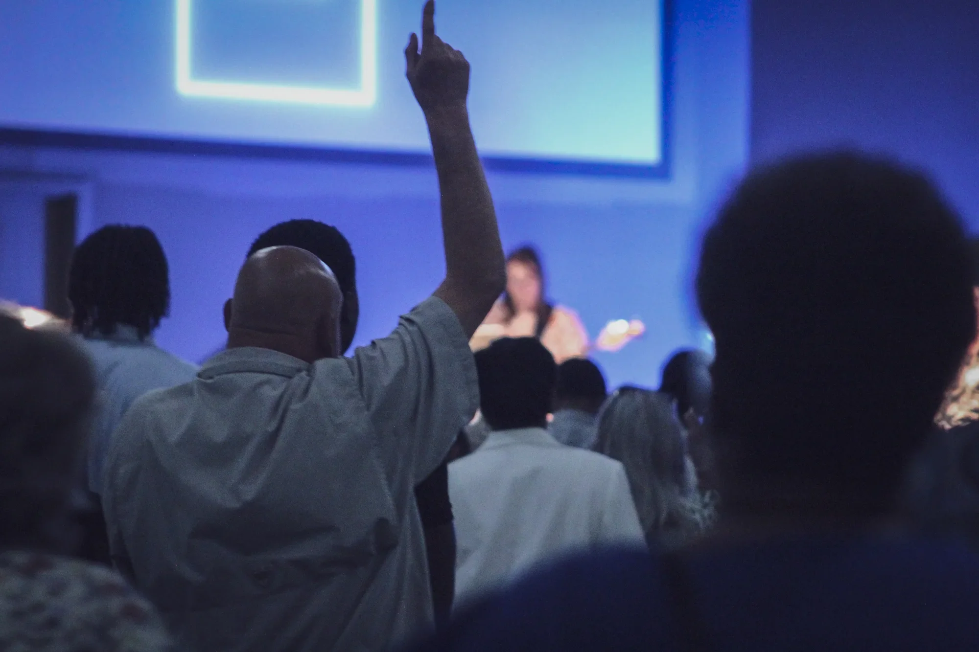 A man in the audience raises his hand in worship during a worship service in a darkened room with a large screen at the front.