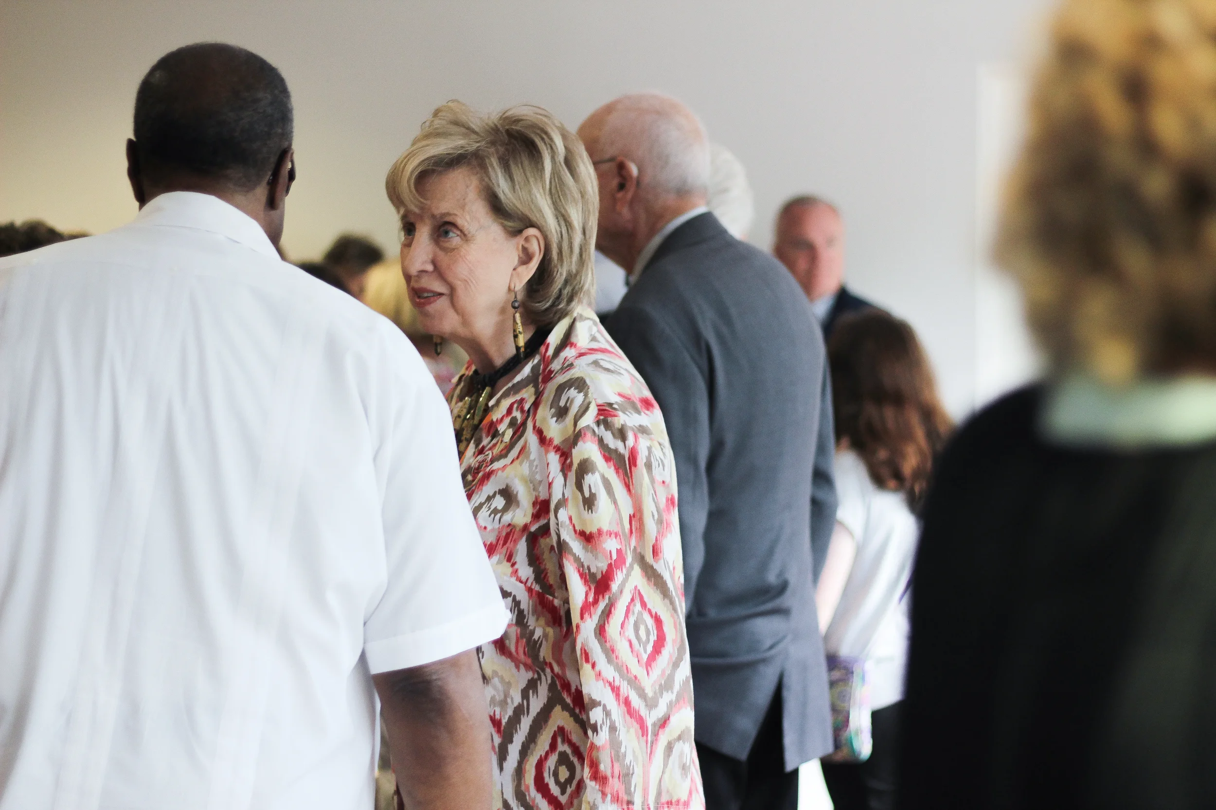 A woman wearing a patterned dress and earrings, talking to a man in a white shirt, at a social gathering with several people in the background.
