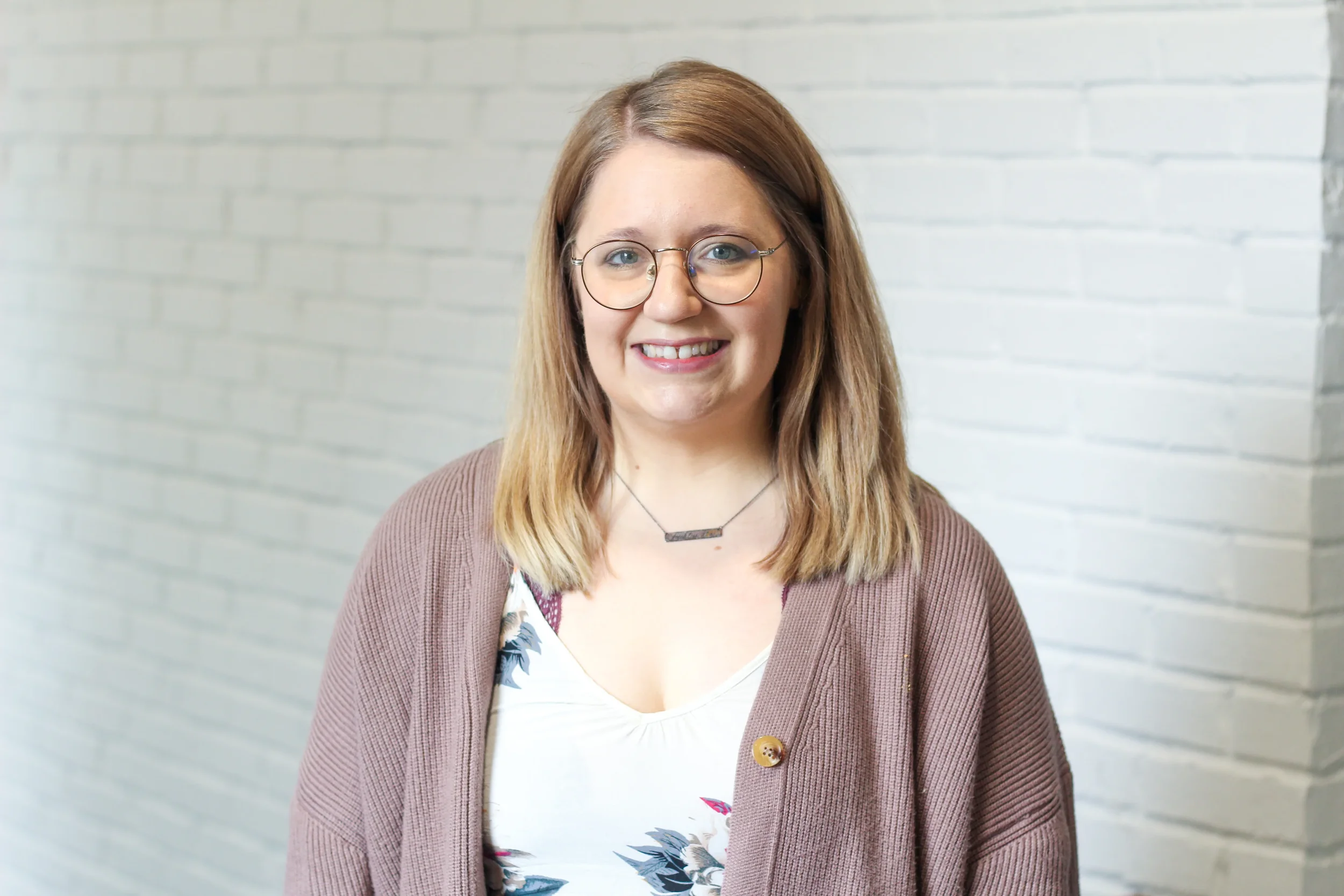 A woman with shoulder-length light brown hair, wearing glasses, a brown cardigan, and a floral top, standing in front of a white brick wall.