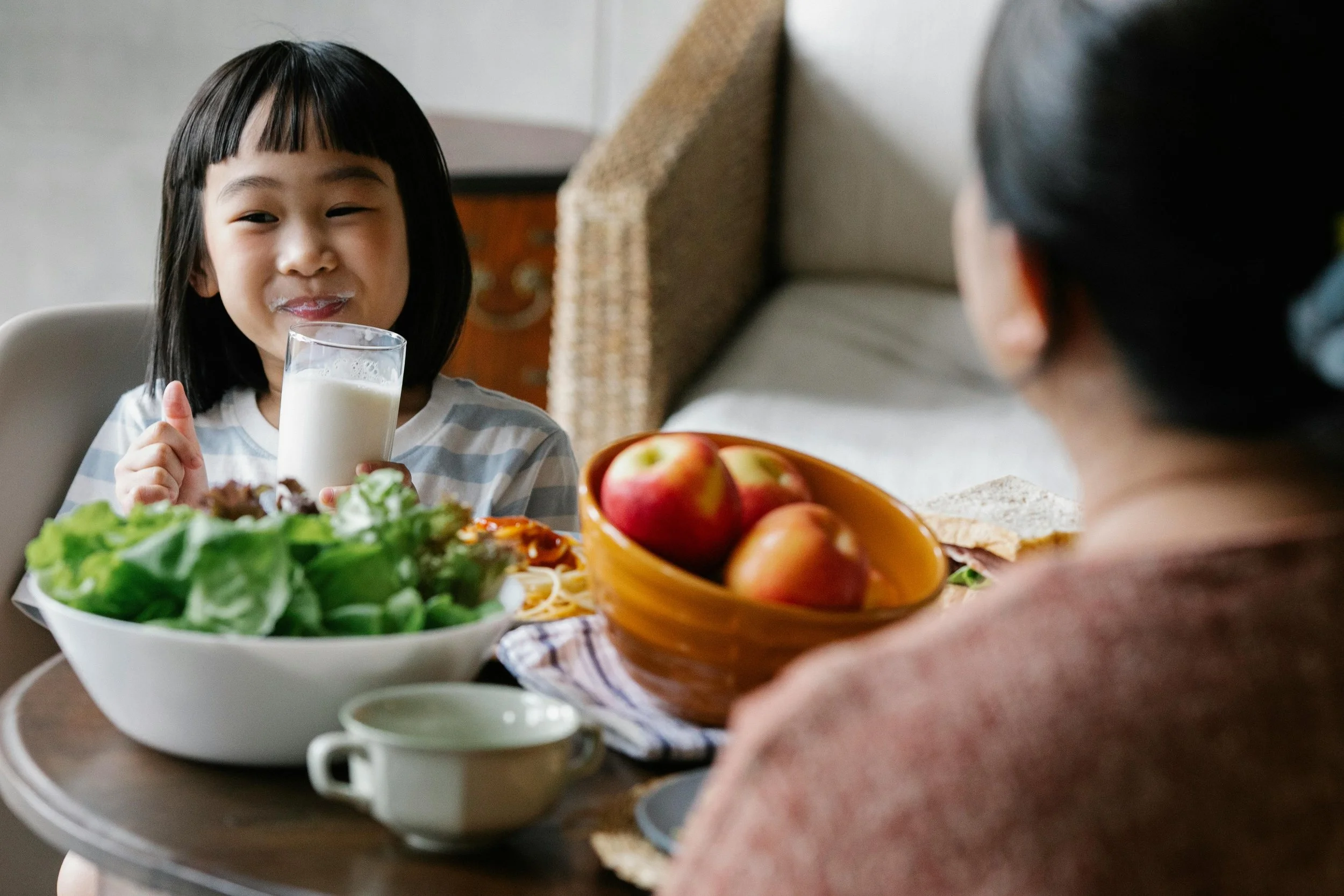 A young girl with dark hair and bangs smiling while holding a glass of milk at a table with a large bowl of salad, a bowl of apples, and other breakfast foods. An older woman is sitting across from her, partially visible.