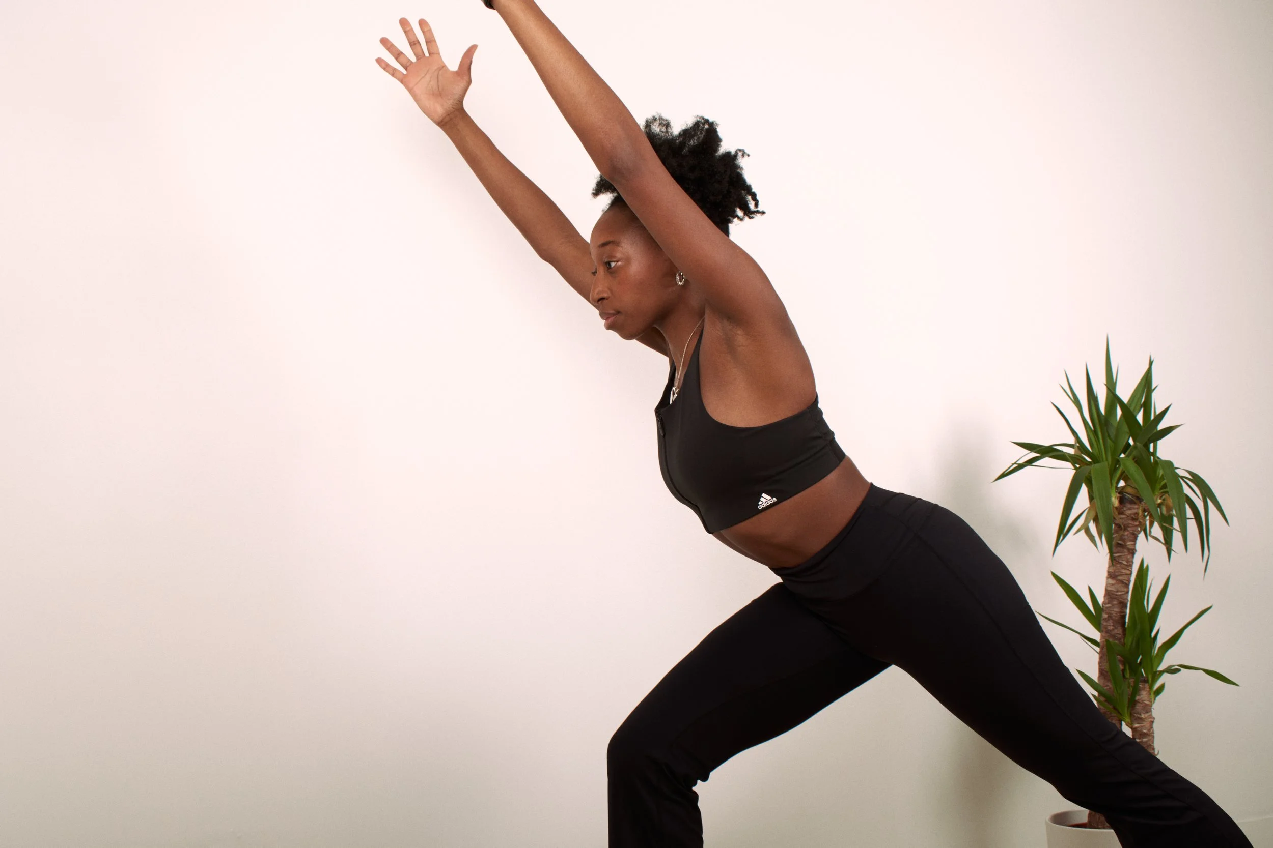 Personne faisant une pose de pilates en tenus de fitness noire, le centre engagé devant un mur blanc et une plante en pot.