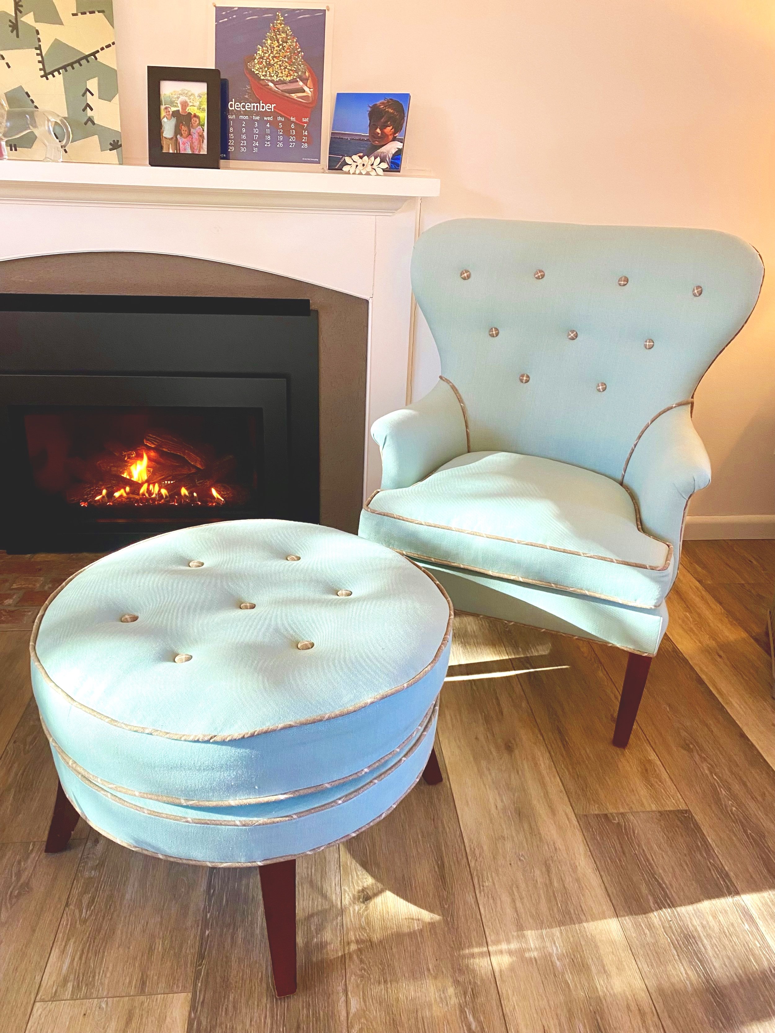 A custom reupholstered light blue linen armchair with a matching round linen ottoman is placed on a wooden floor in front of a fireplace. The fireplace has a lit fire. Eco-friendly upholstery. Brewster, MA. Cape Cod Upholstery