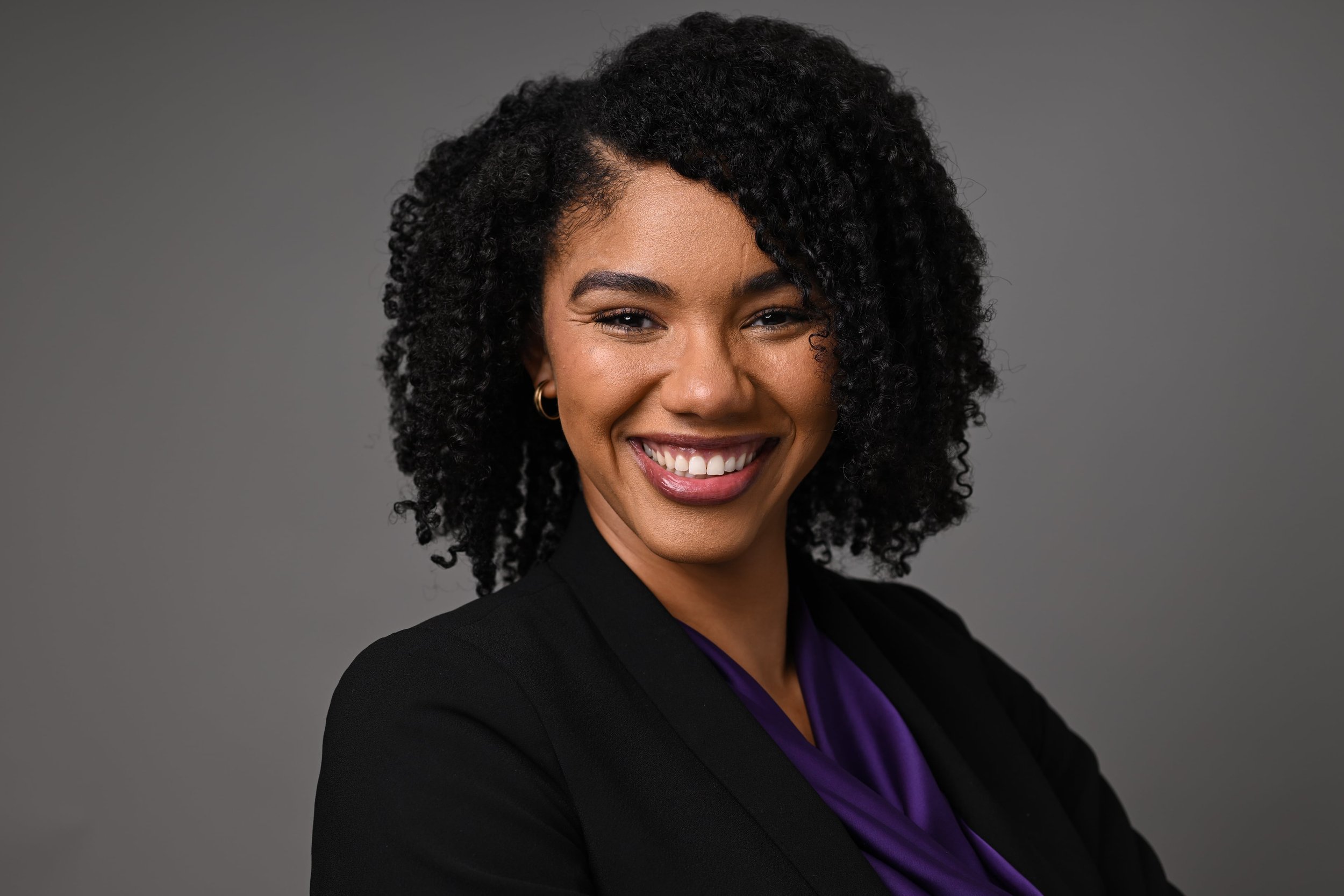Smiling person with curly hair in a black blazer, sitting on a desk with arms crossed, in a professional office setting.