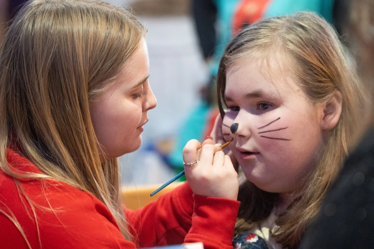 Two girls, one painting whiskers and a nose on the other's face, using face paint and a brush.