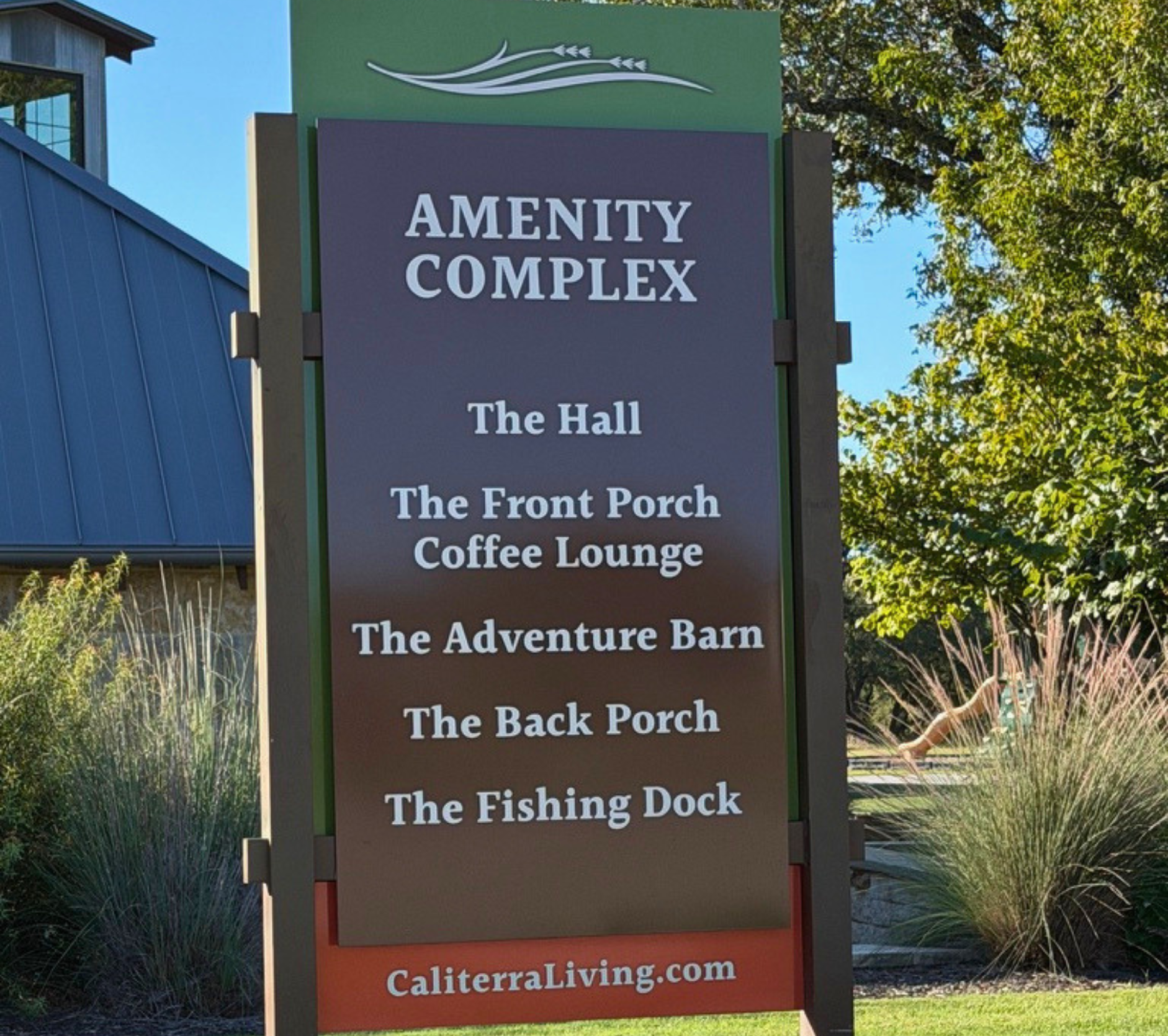 A tall signboard with white lettering on a brown background listing amenities: The Hall, The Front Porch Coffee Lounge, The Adventure Barn, The Back Porch, The Fishing Dock. The top of the sign features a stylized graphic of flowing lines with arrowh