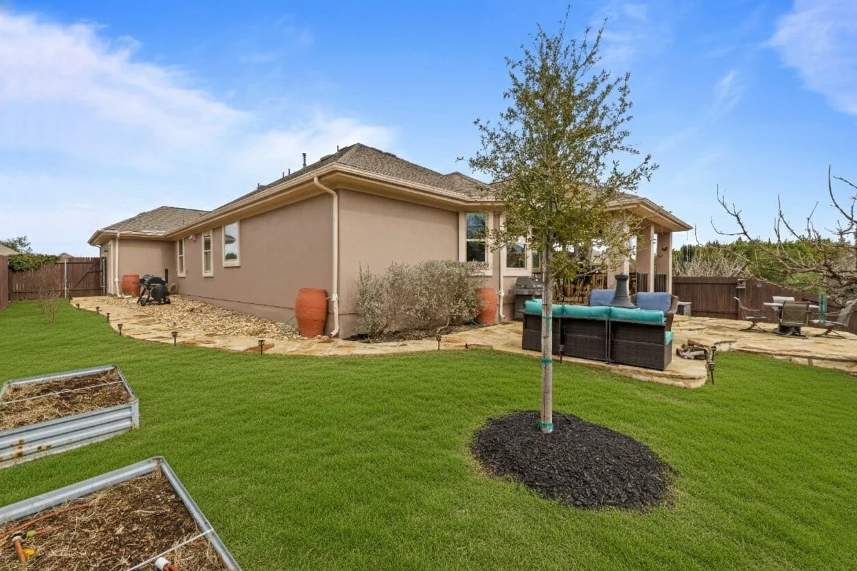 Backyard of a house with a lawn, young tree, patio furniture, and garden beds.