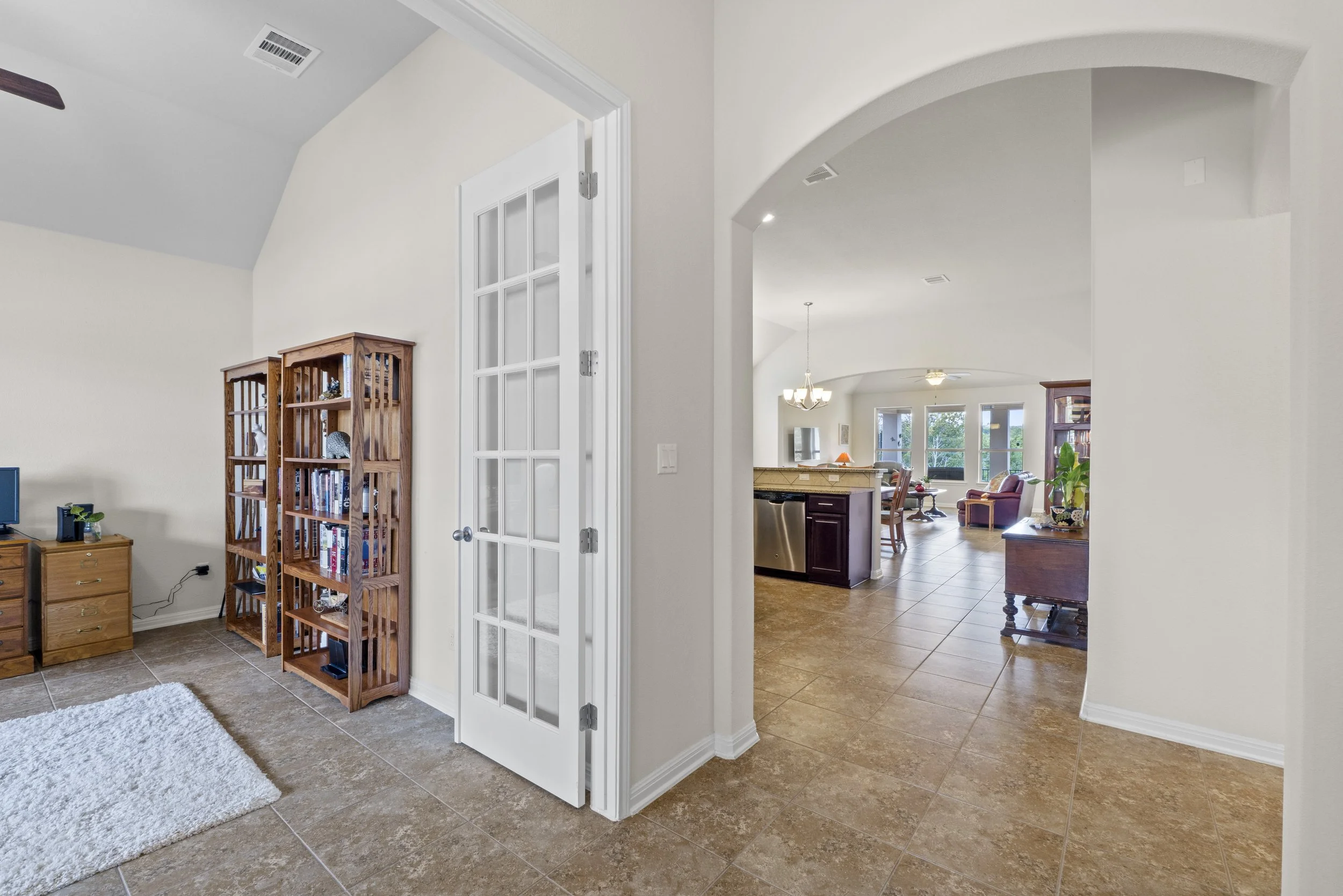 Interior view of a house showing a living room and kitchen with tile flooring, white walls, and large windows.