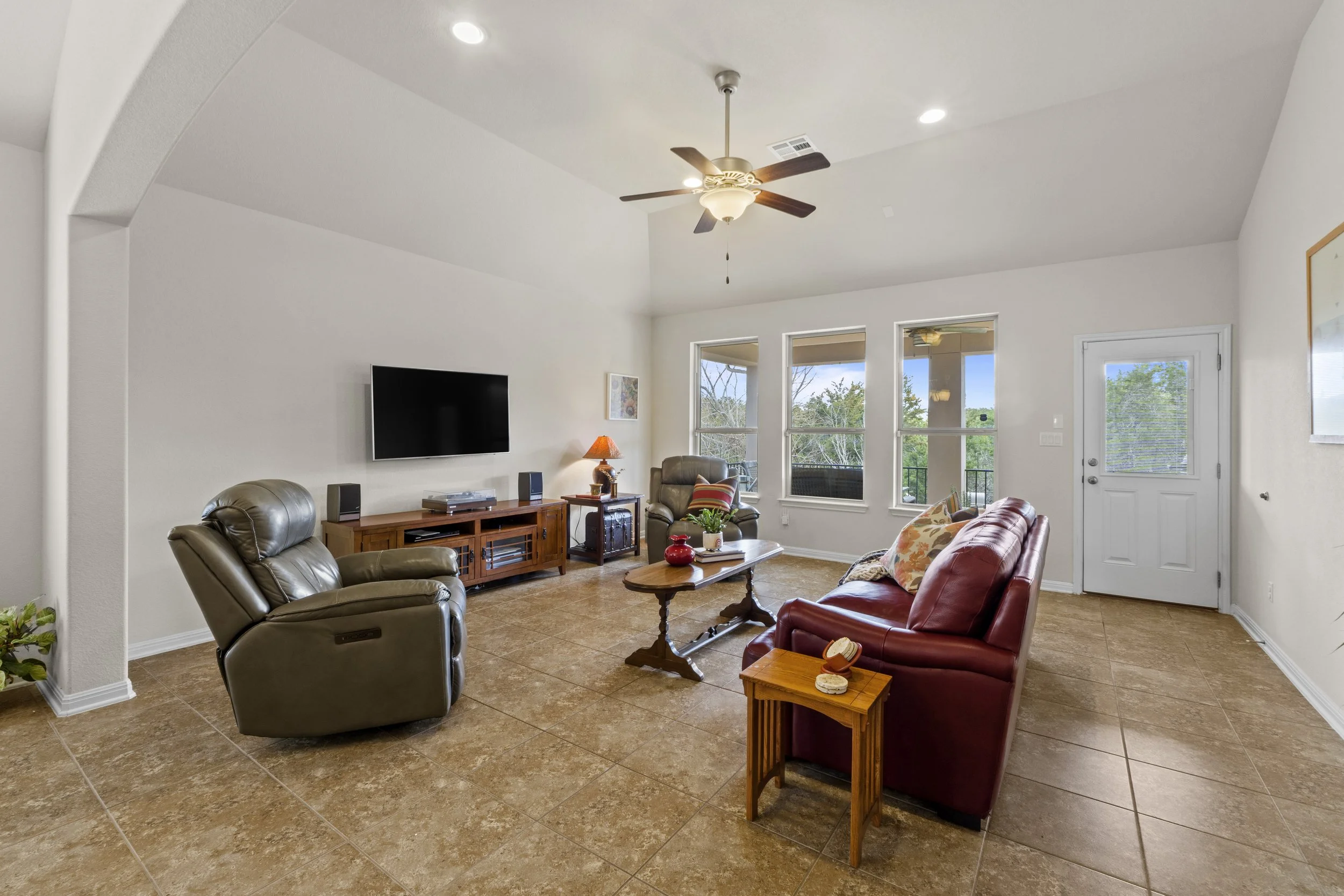 Living room with leather chairs, a red sofa, and a wooden coffee table. Large windows providing natural light, ceiling fan, and a wall-mounted TV.