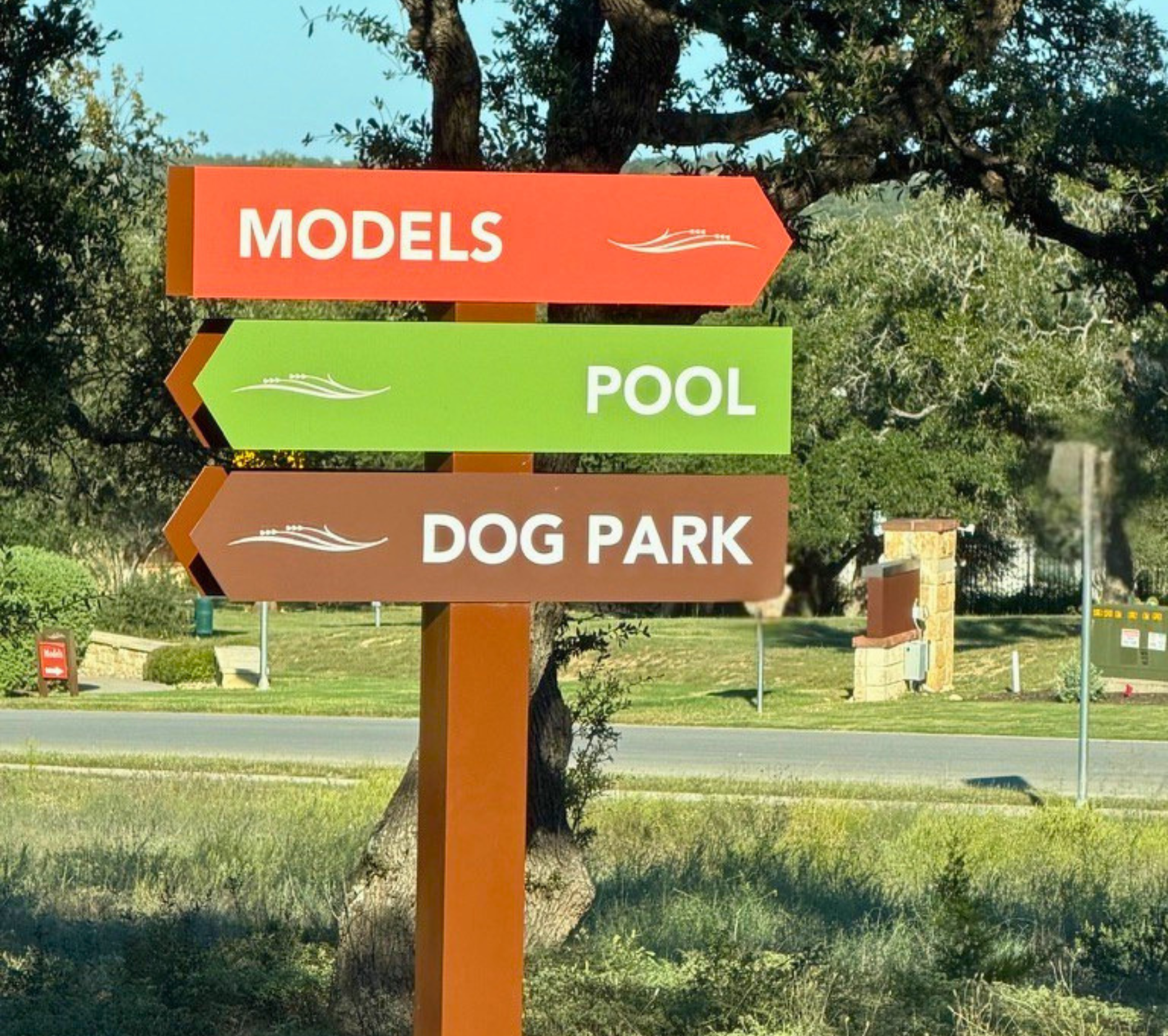 Colorful directional signpost with arrows pointing right and left, indicating directions to models, pool, and dog park, set outdoors with trees and grass in the background.