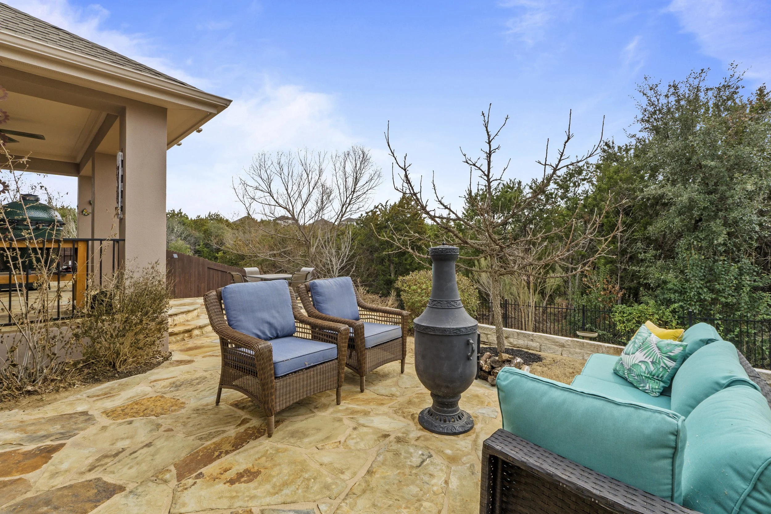 Outdoor patio with rattan loveseat with blue cushions, a teal outdoor sofa with decorative pillows, a black chiminea, and a view of trees with bare branches and green foliage under a blue sky.