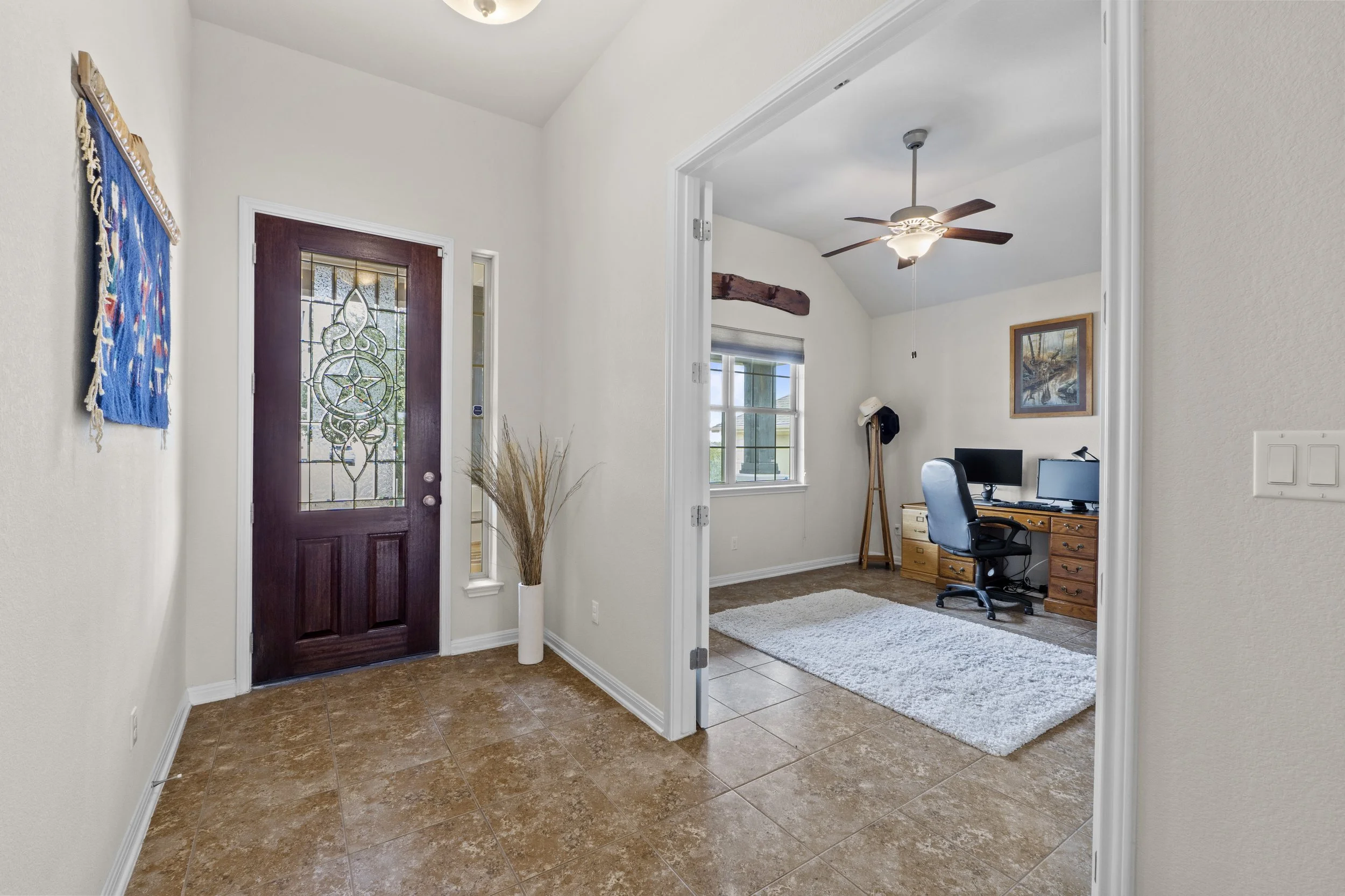 Entryway with a wooden front door with stained glass, a white wall with artwork, and a tall white vase with dried grasses. An opening leads to a home office with a desk, monitor, desk lamp, computer chair, window, and ceiling fan.