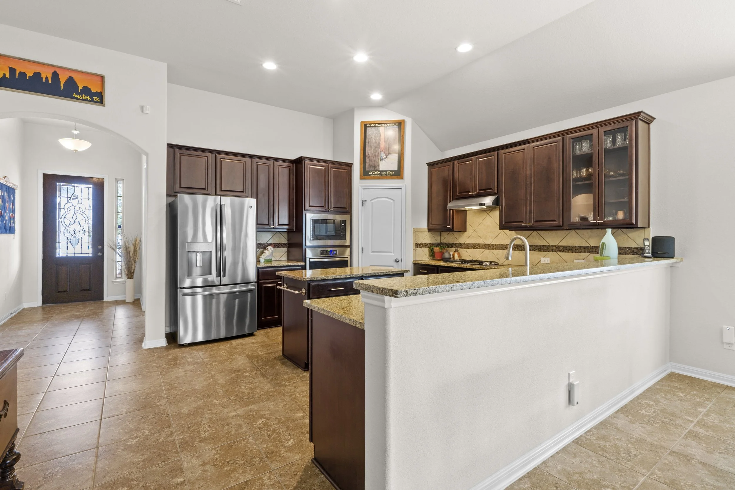 Modern kitchen with stainless steel refrigerator, dark wood cabinets, beige granite countertops, tiled backsplash, and an open counter area for dining or serving.