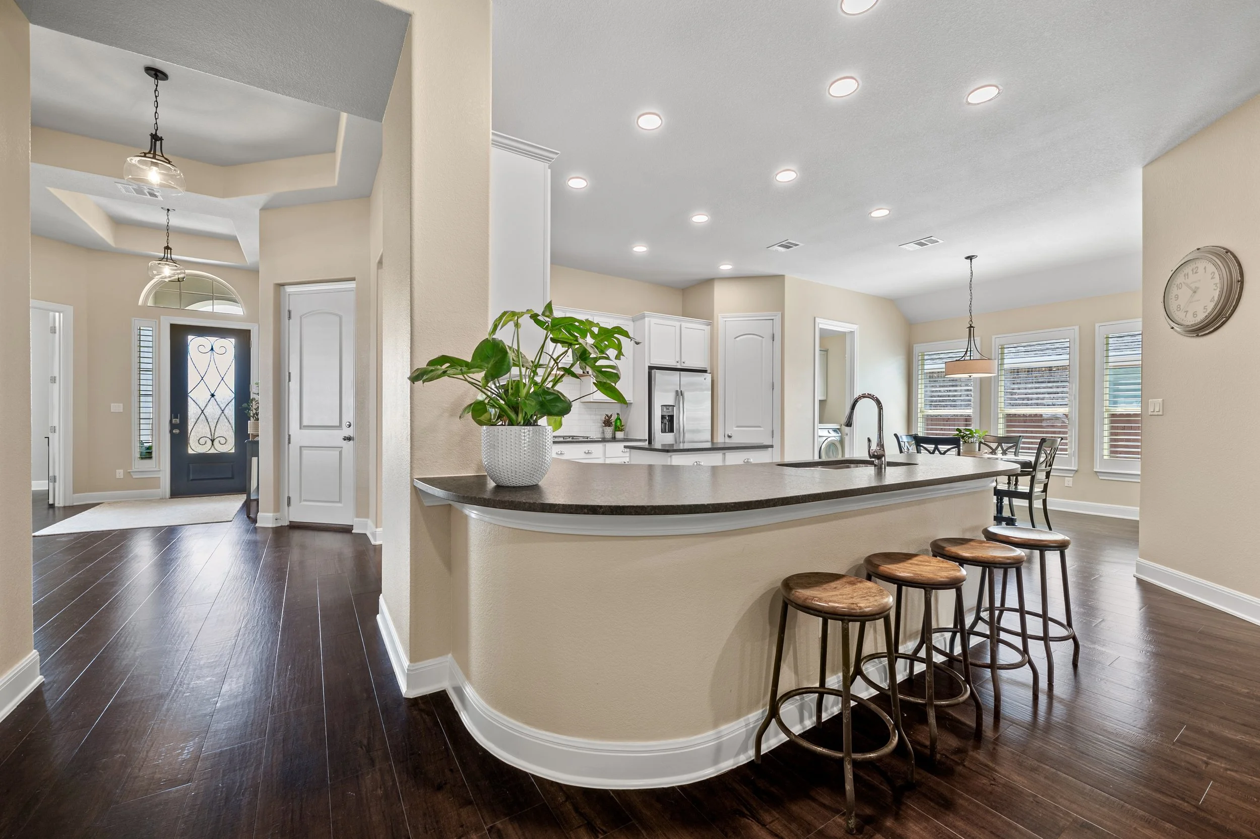 Open-concept kitchen with a curved island, white cabinets, stainless steel refrigerator, and dining area with a round table and chairs. Dark hardwood floors, beige walls, multiple windows, and potted plant on the island.