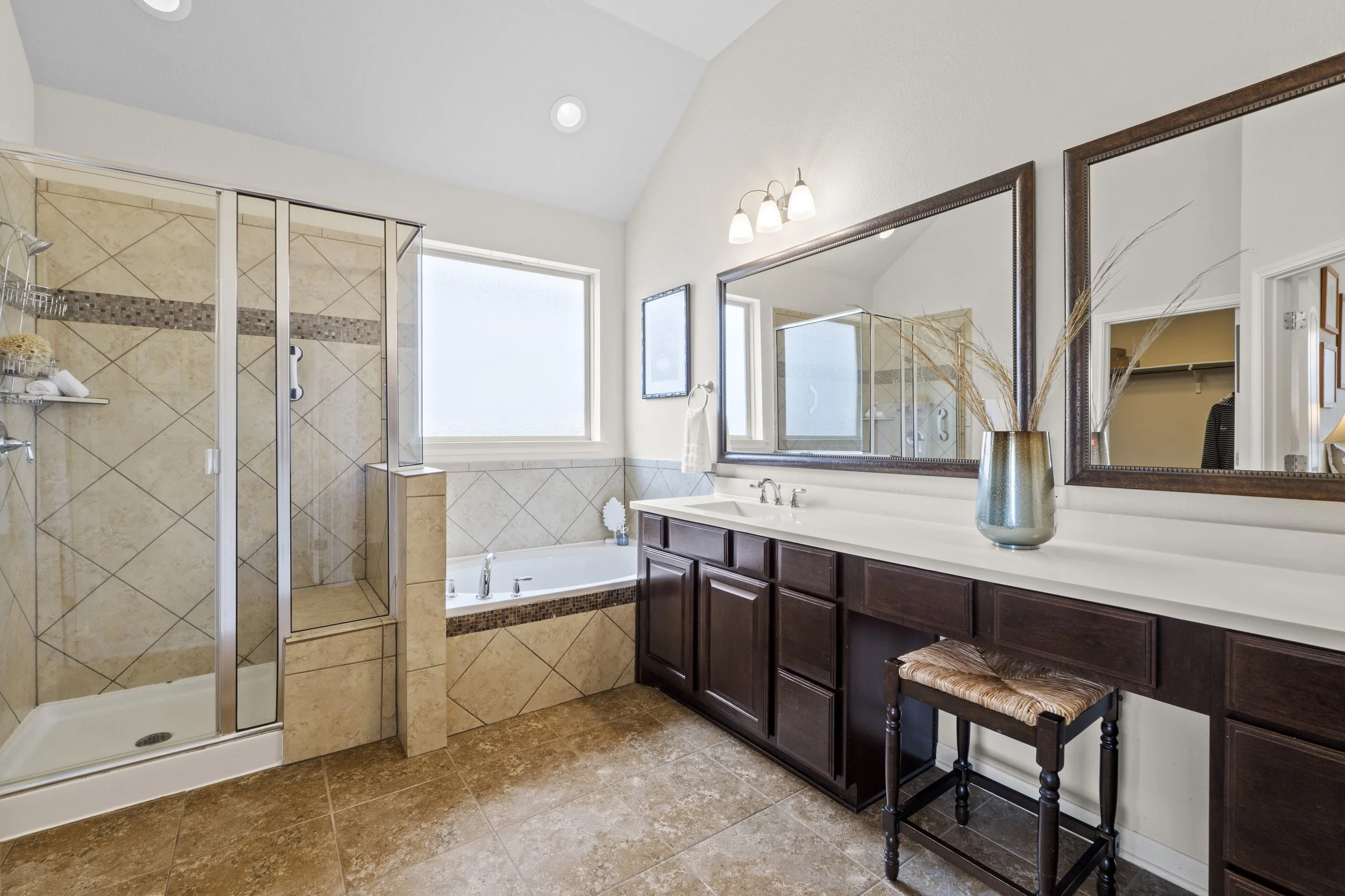 A spacious bathroom with a walk-in shower, a bathtub beneath a frosted window, a large mirror above a dark wooden vanity, and a decorative vase with dried plants on the counter.