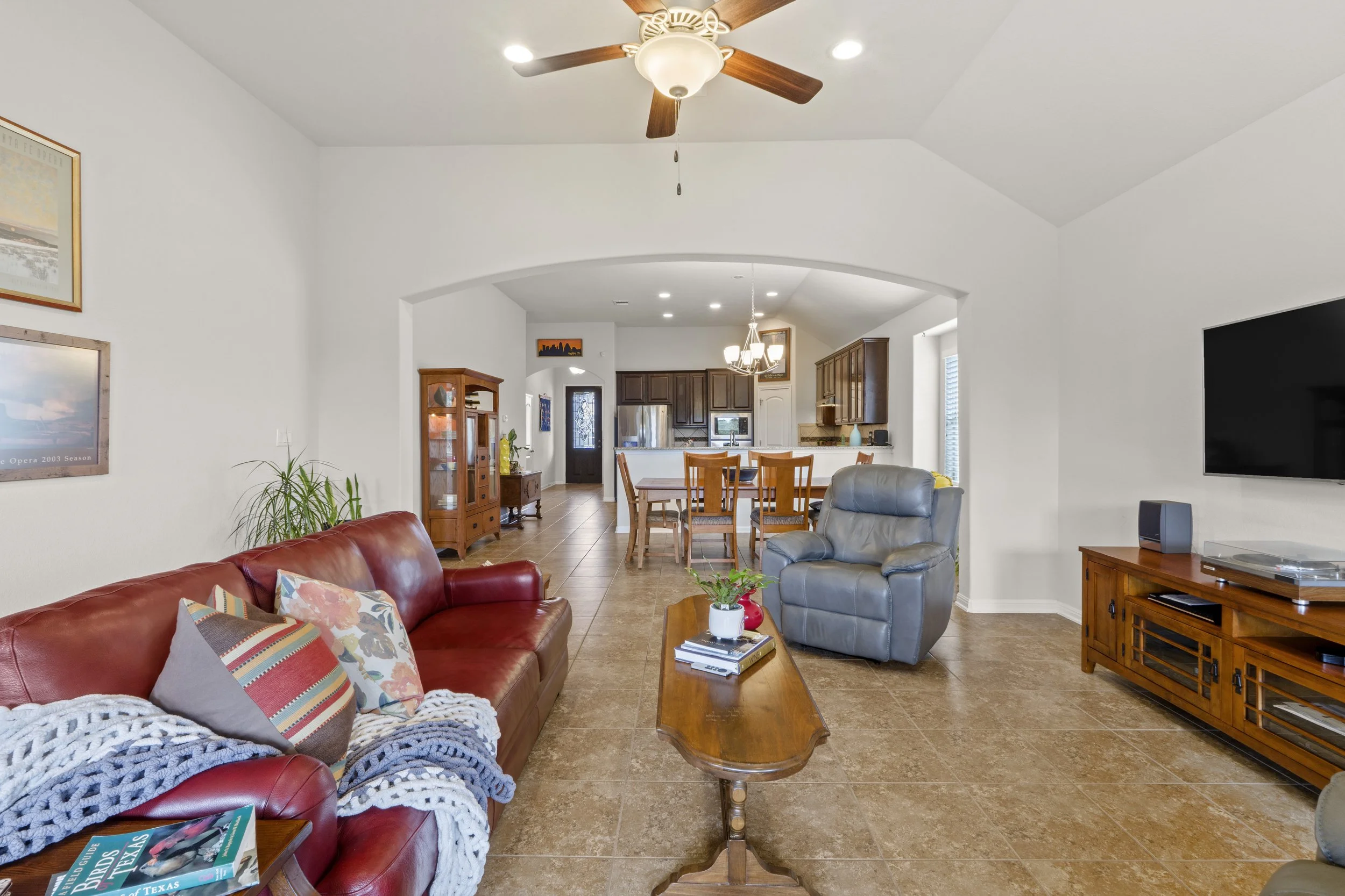 Living room with a red leather sofa, a gray recliner, a wooden coffee table with plants, and a large flat-screen TV on a wooden stand. The room opens to a dining area and kitchen with wooden cabinets and a chandelier.