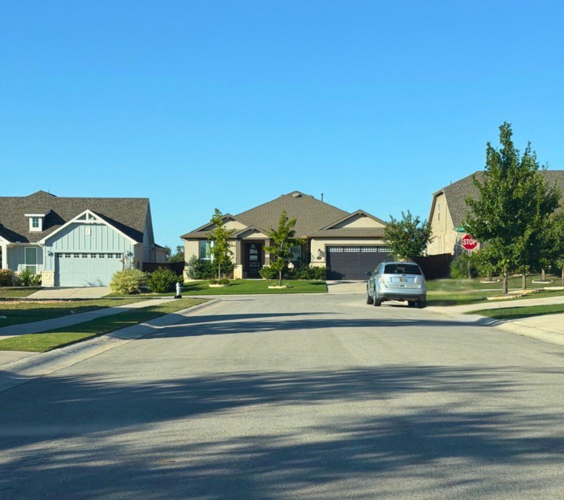 A suburban neighborhood street scene featuring several houses with garages and driveways, a silver sedan parked, a stop sign, green trees, and a clear blue sky.