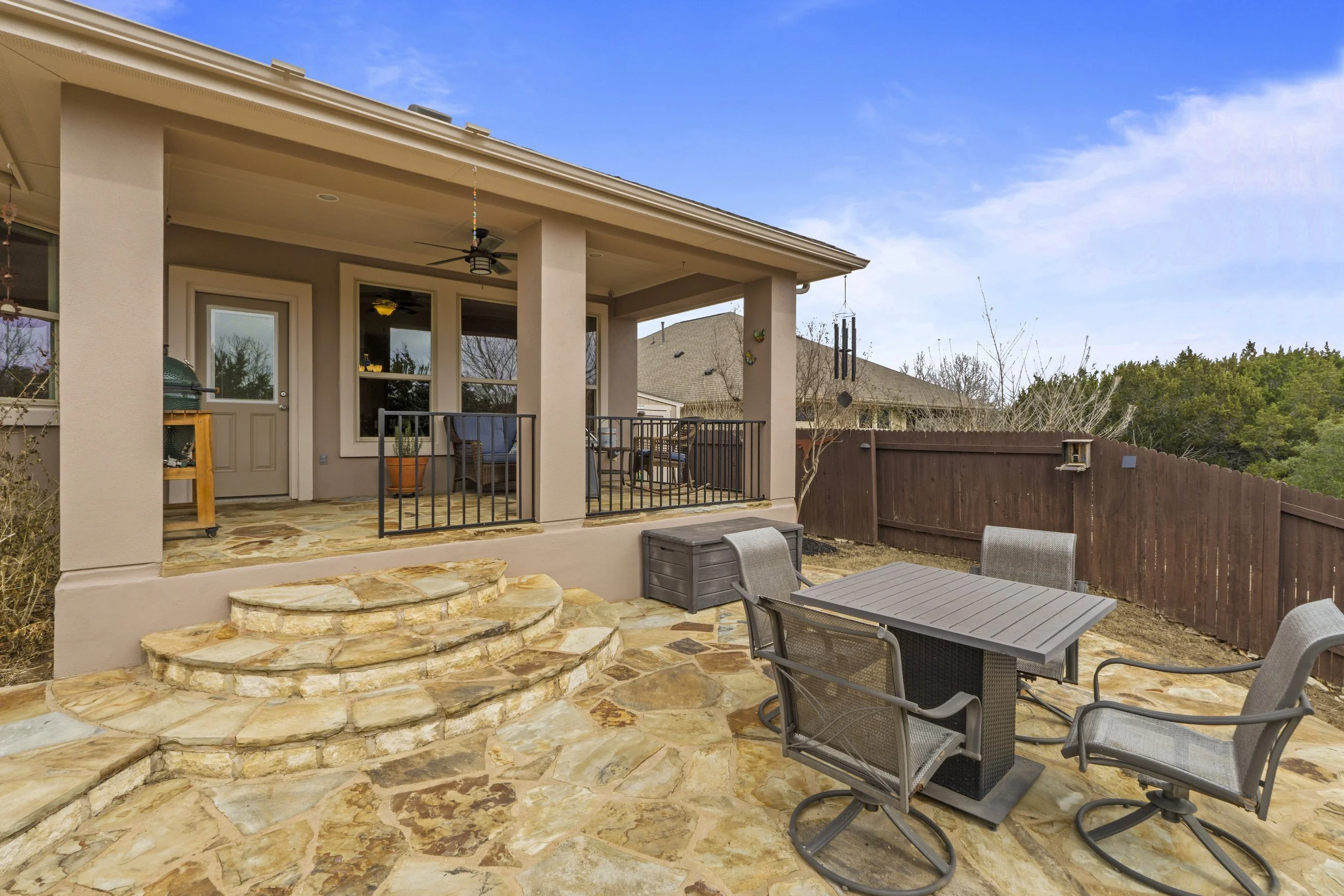 Backyard patio with stone steps leading up to a covered porch, outdoor dining table with four chairs, enclosed by a wooden fence, with trees in the background, and a partly cloudy sky.
