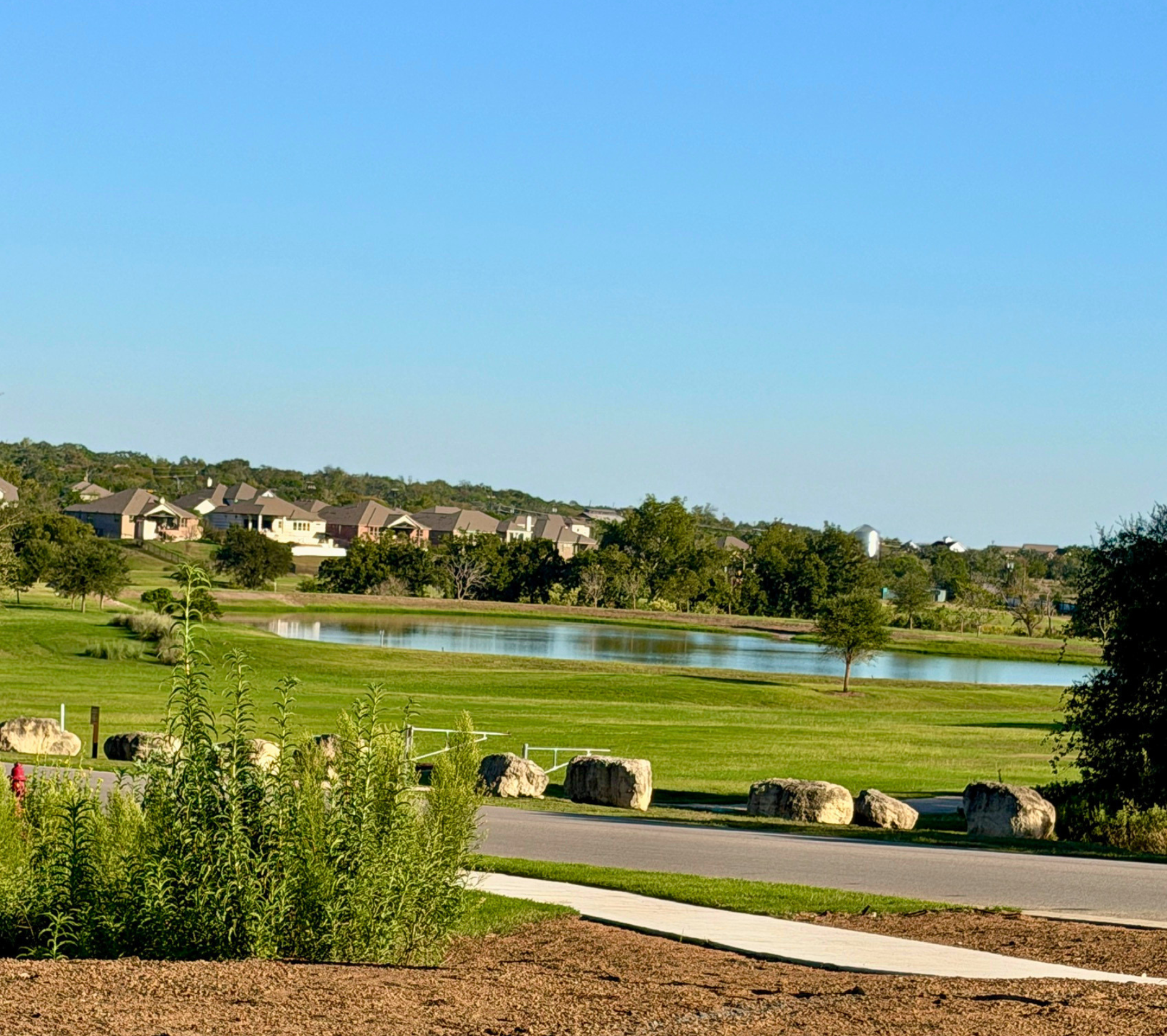 A suburban neighborhood with rows of modern houses on a hill, a pond, green lawns, trees, and a clear blue sky.